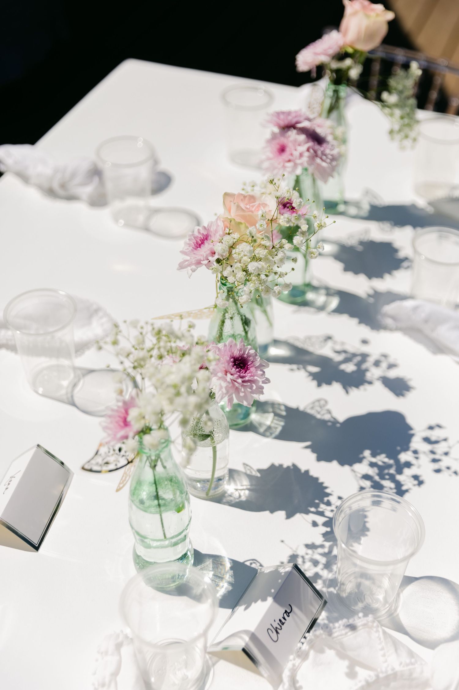 Sunlit reception table with delicate floral arrangements in glass bottles casting dramatic shadows.