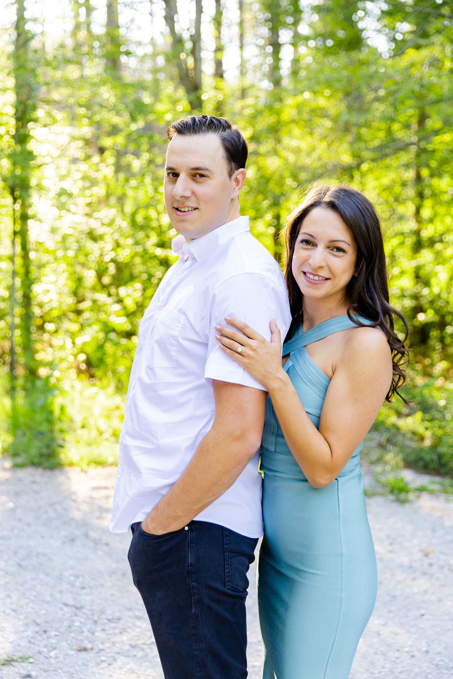 A couple poses together on a sunlit forest path with the woman in a blue dress and man in white shirt.
