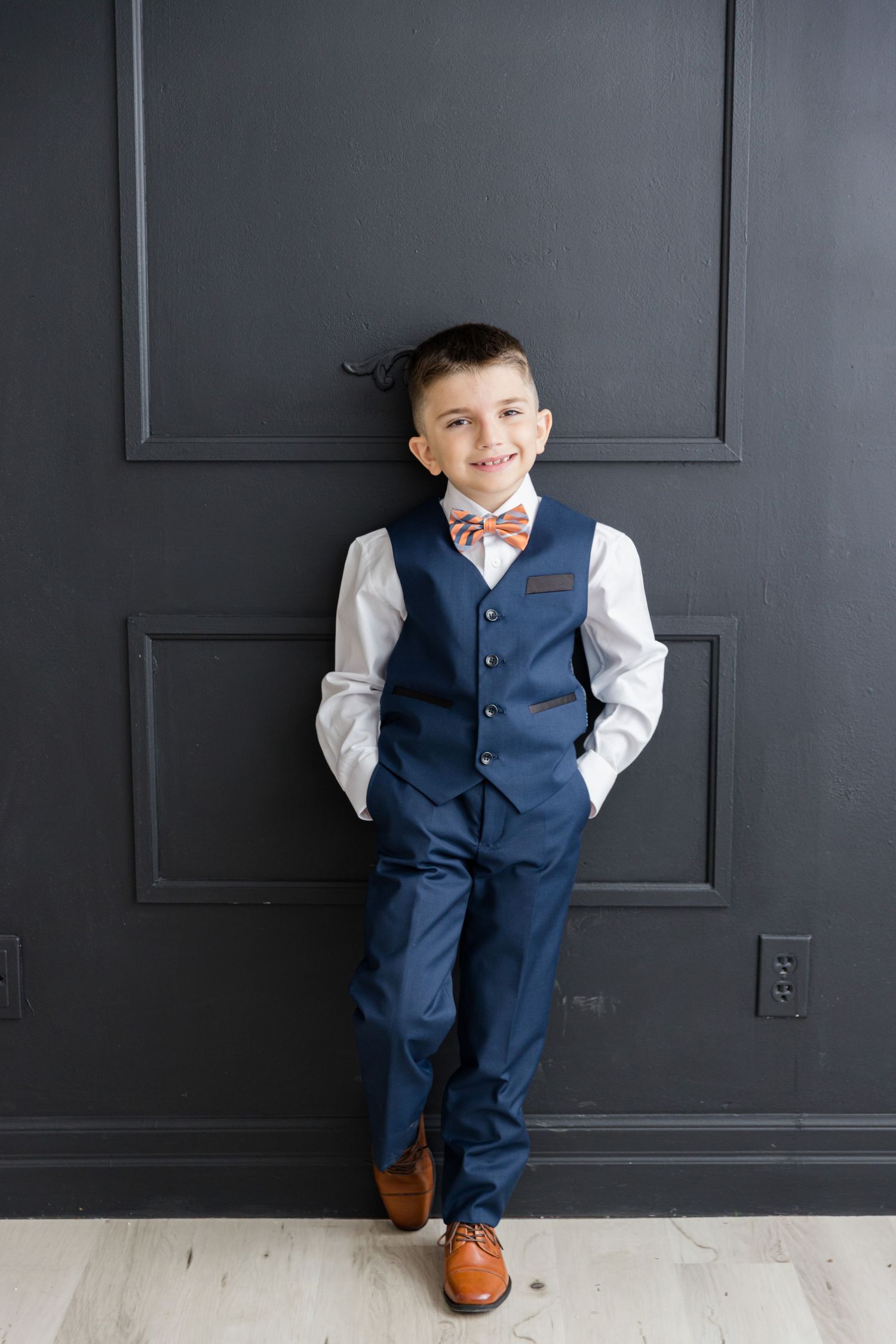 Formal outfit with navy vest and bow tie standing against dark paneled wall with brown shoes.