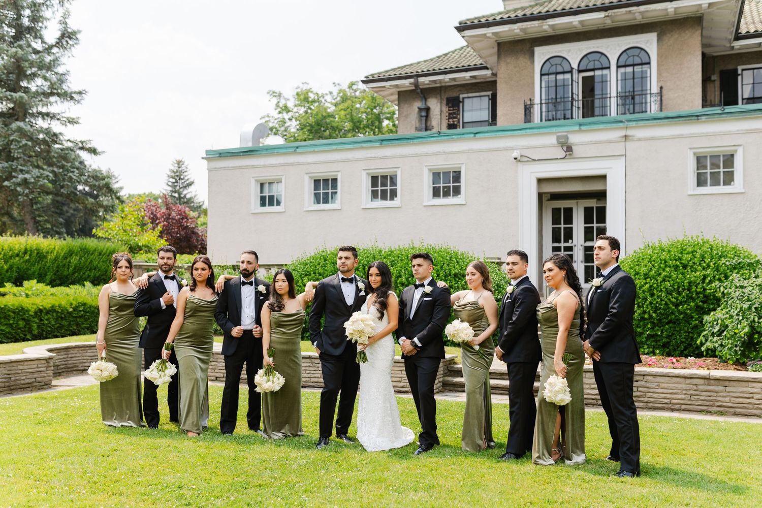 Full wedding party with bridesmaids in sage and groomsmen in black lined up on mansion lawn.