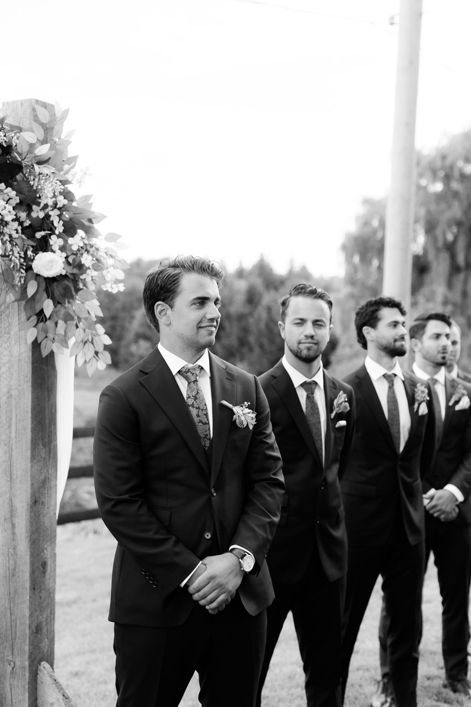 Black and white photo of wedding party members in formal attire standing at outdoor ceremony.