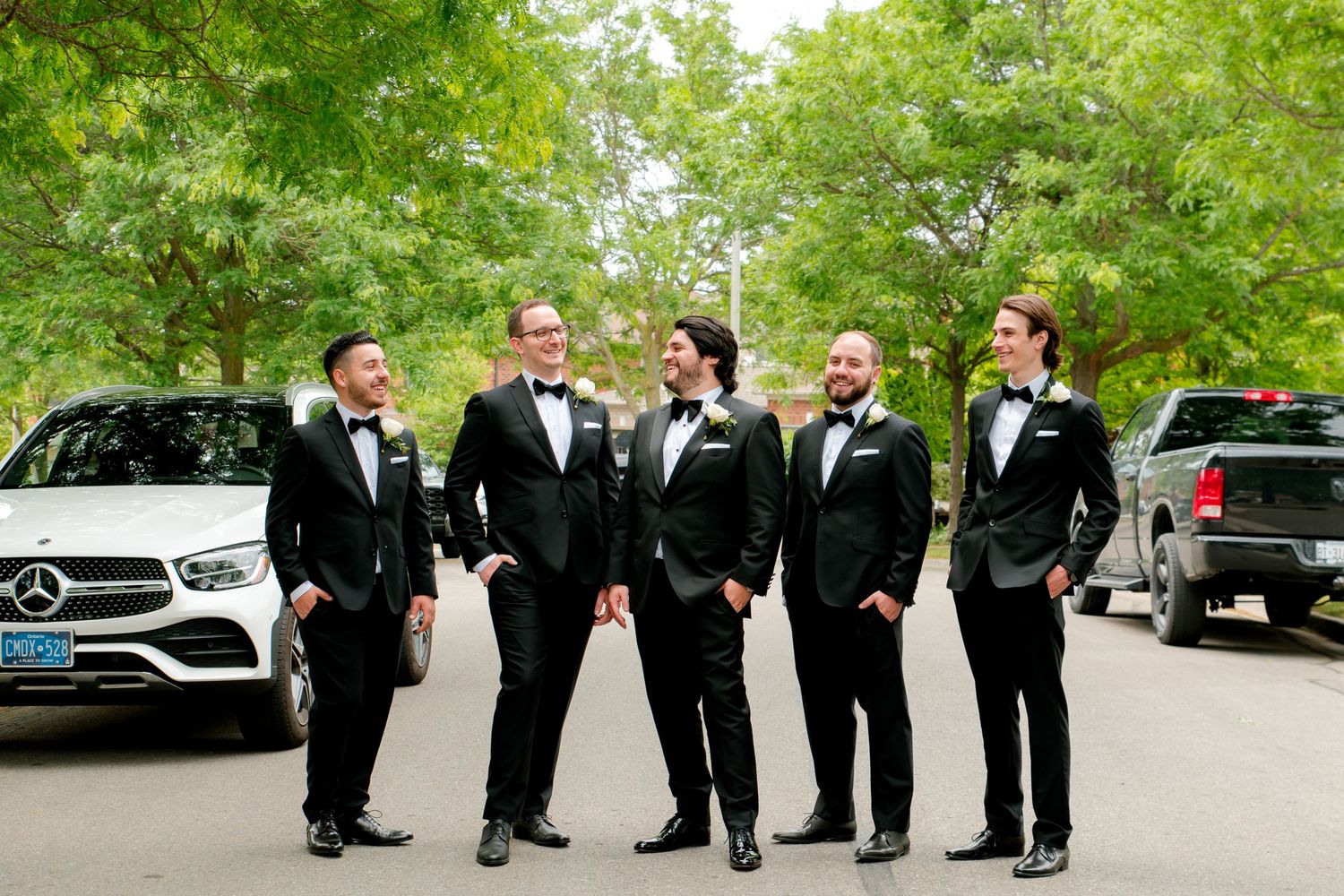 Five men in black tuxedos pose on street with luxury cars in background.