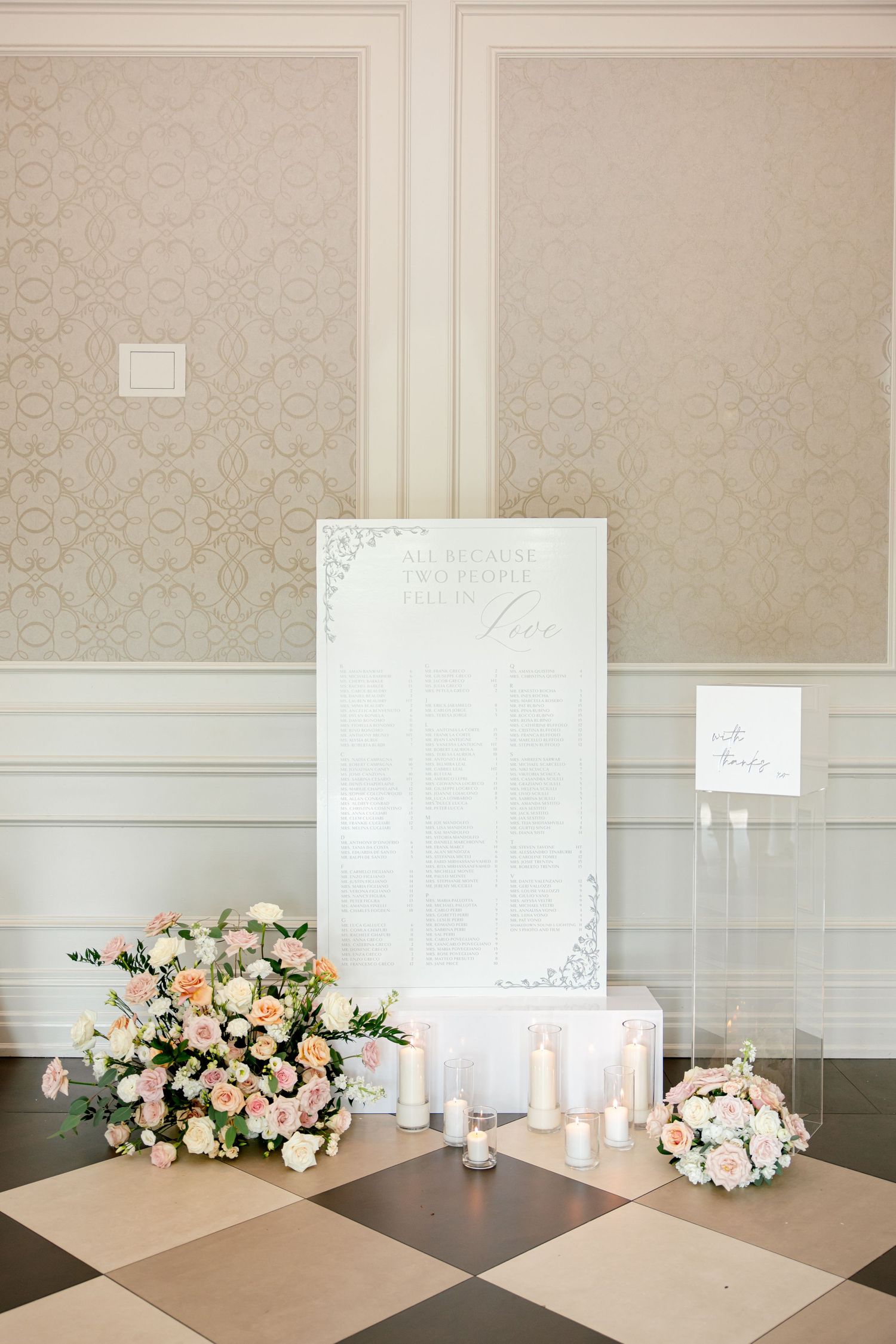 Elegant wedding seating chart display with white board, candles and peach floral arrangements on checkered floor.