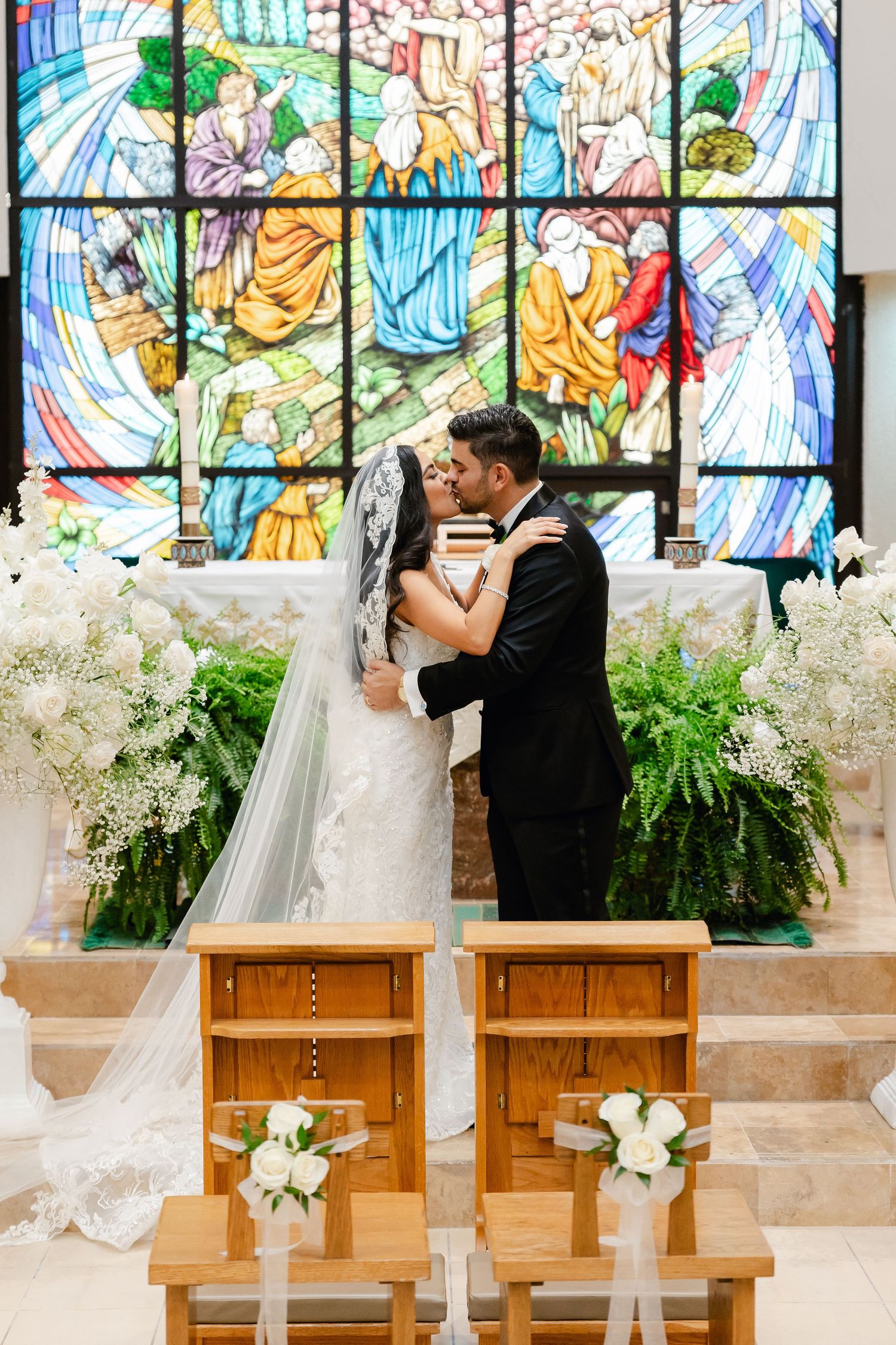 A wedding ceremony in front of a vibrant stained glass window with white floral decorations in a church.