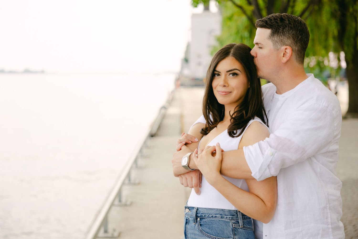 A couple poses together by the water, with trees in the background.