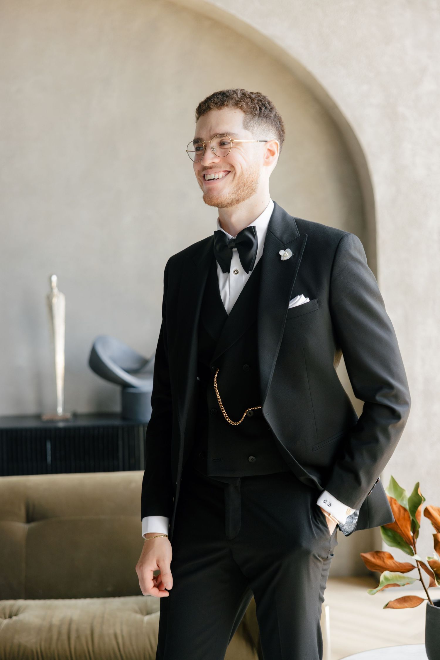 Groom in black tuxedo and bow tie smiles warmly while standing in an elegant room.
