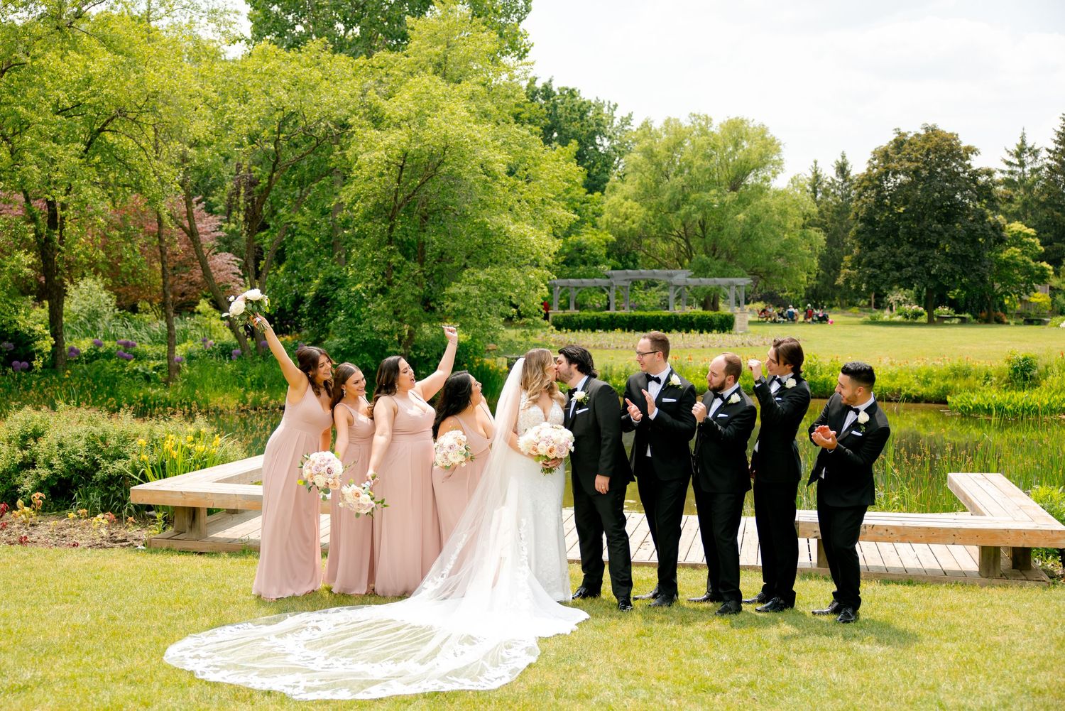 Wedding party celebrates joyfully on wooden walkway surrounded by greenery with bridesmaids in pink dresses.