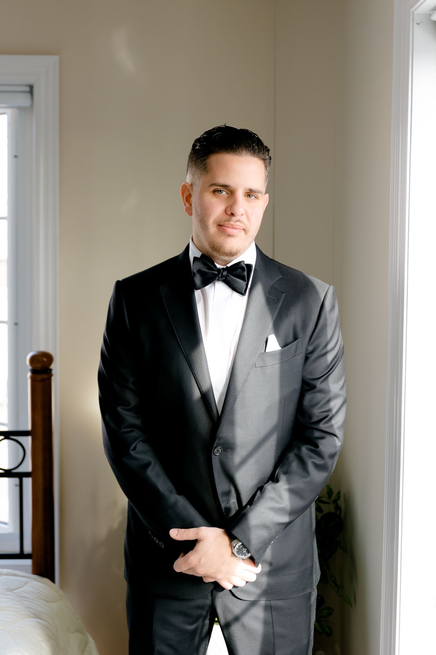 Groom poses in classic black tuxedo with bowtie against neutral wall.