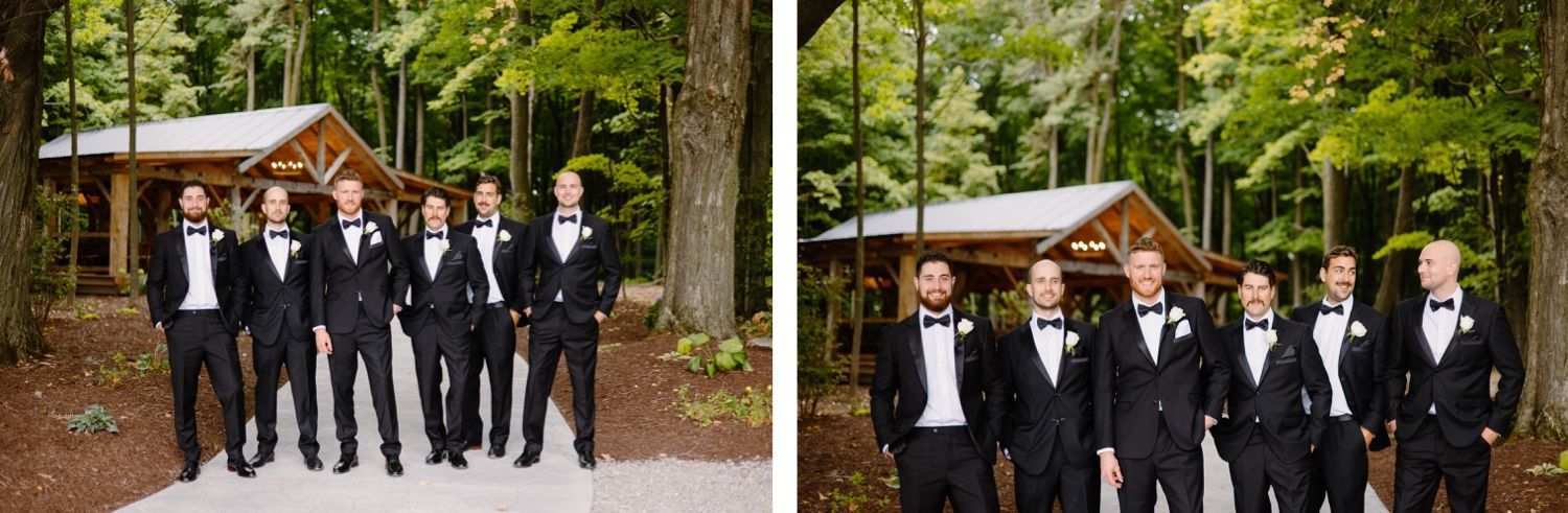 Wedding party members in black tuxedos pose together in wooded outdoor setting.