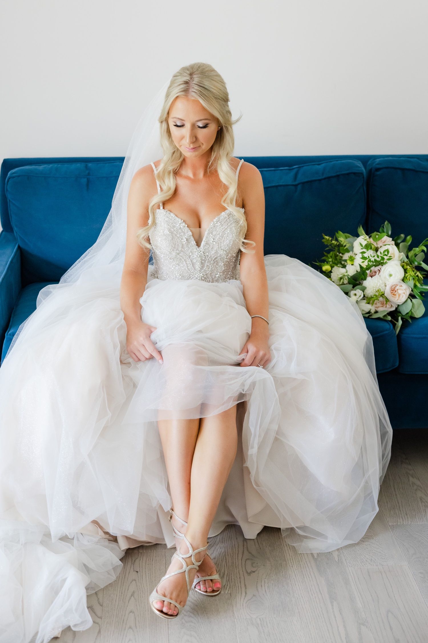 Bride in strapless lace gown showcases her wedding shoes while seated on blue velvet couch.