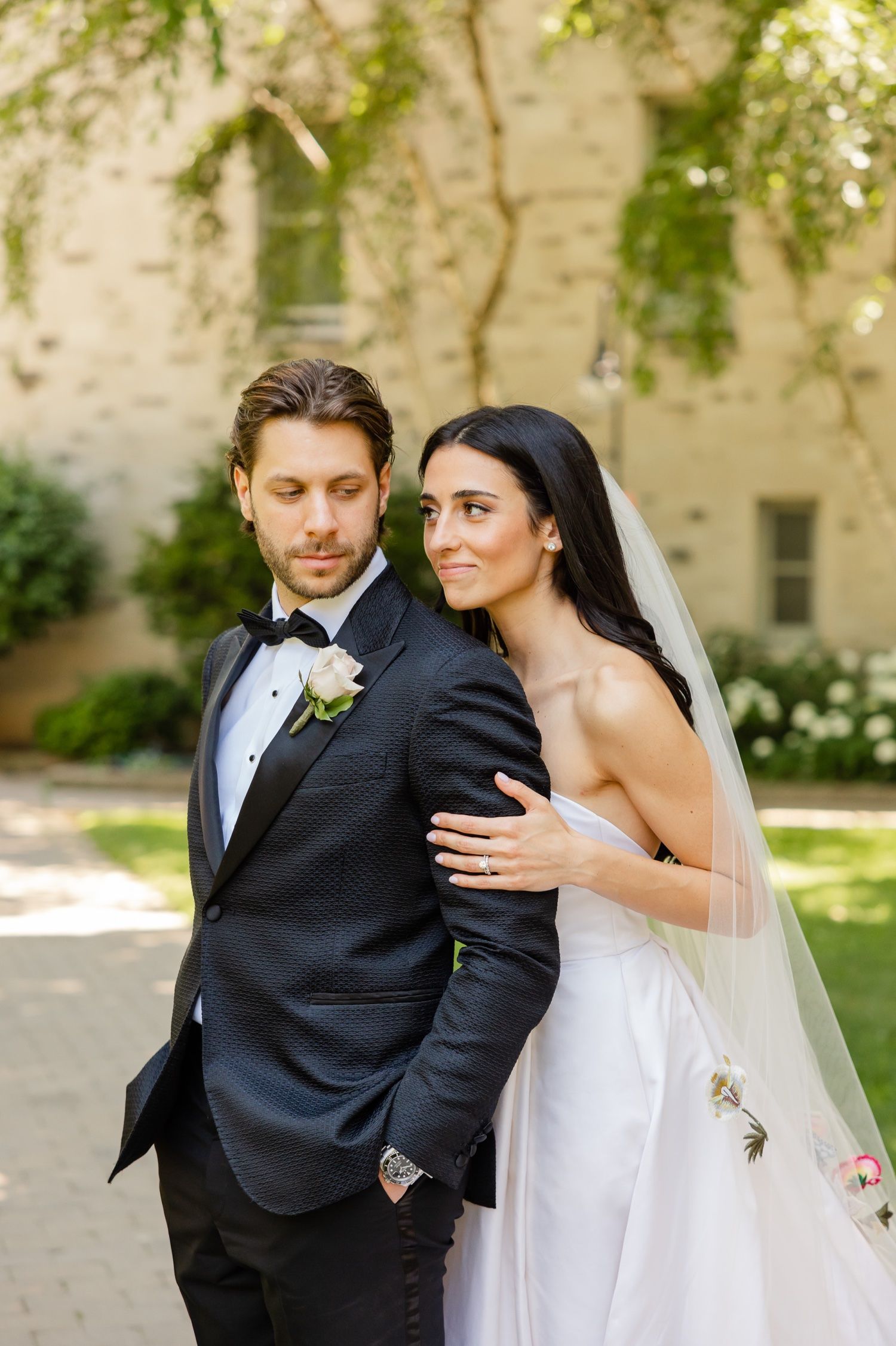 Elegant wedding couple poses outdoors with sunlit stone building and greenery in background.