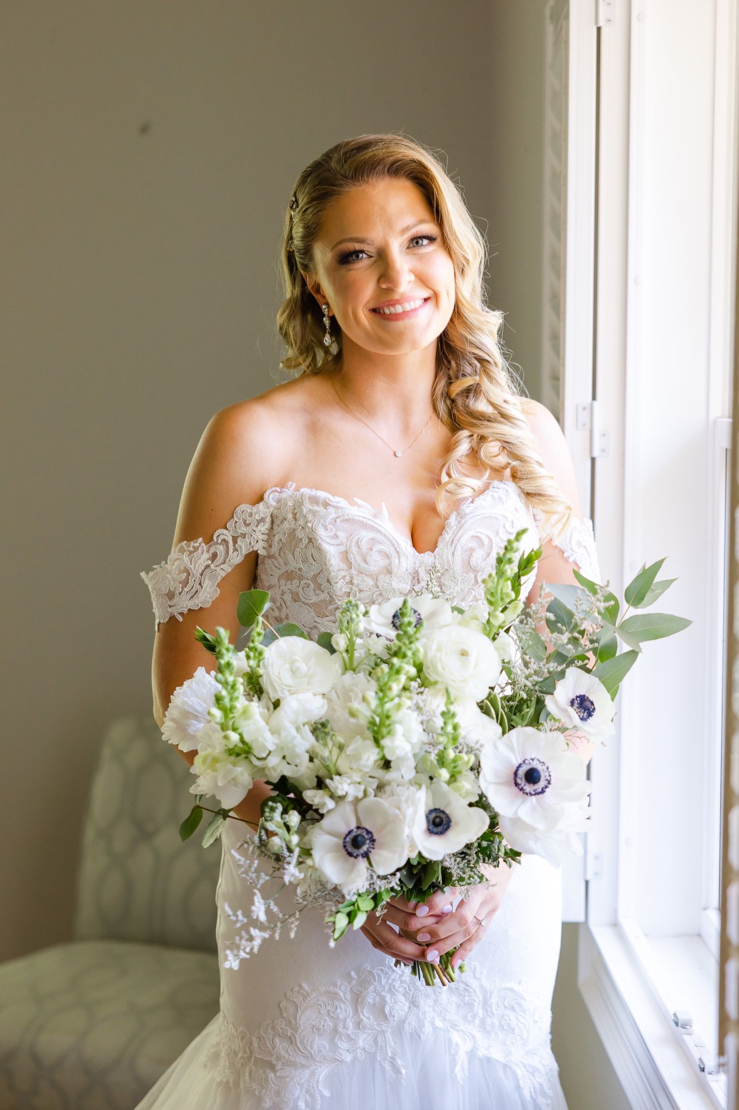 A beautiful bride in a lace off-shoulder wedding gown holds a stunning white and purple anemone flower bouquet.