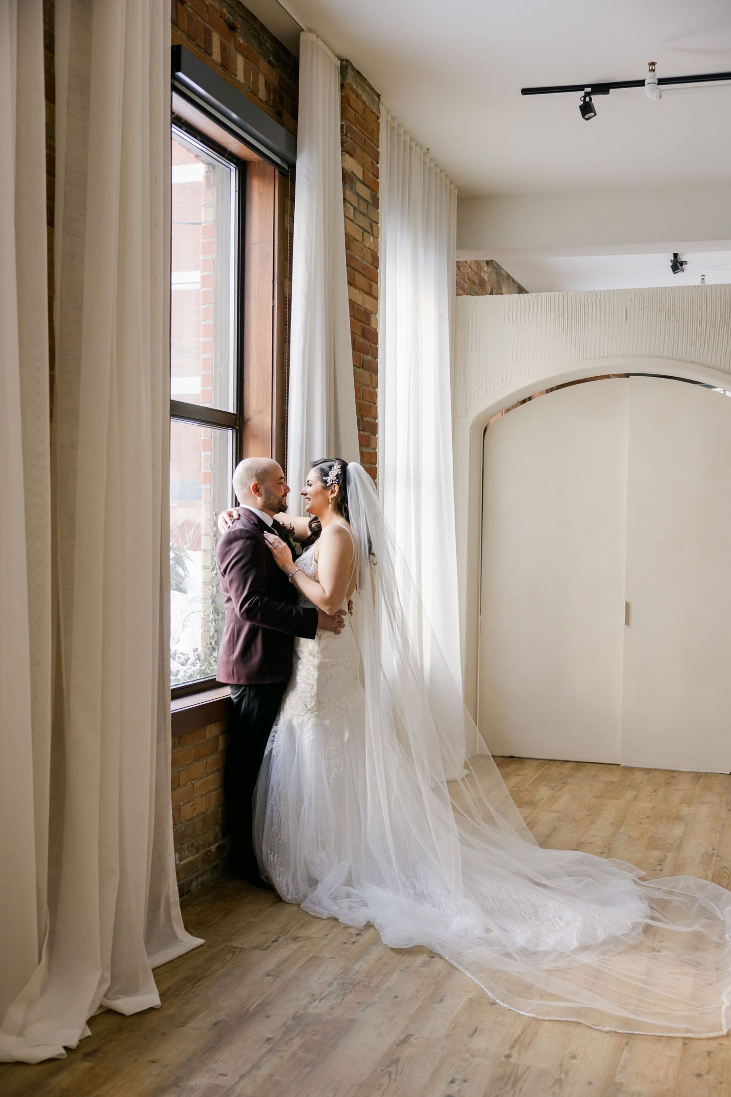 A wedding couple shares an intimate moment near a sunlit window in an elegant venue.