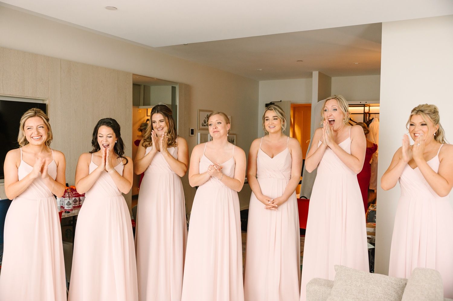 Bridesmaids in blush pink dresses react with excitement during wedding preparations.