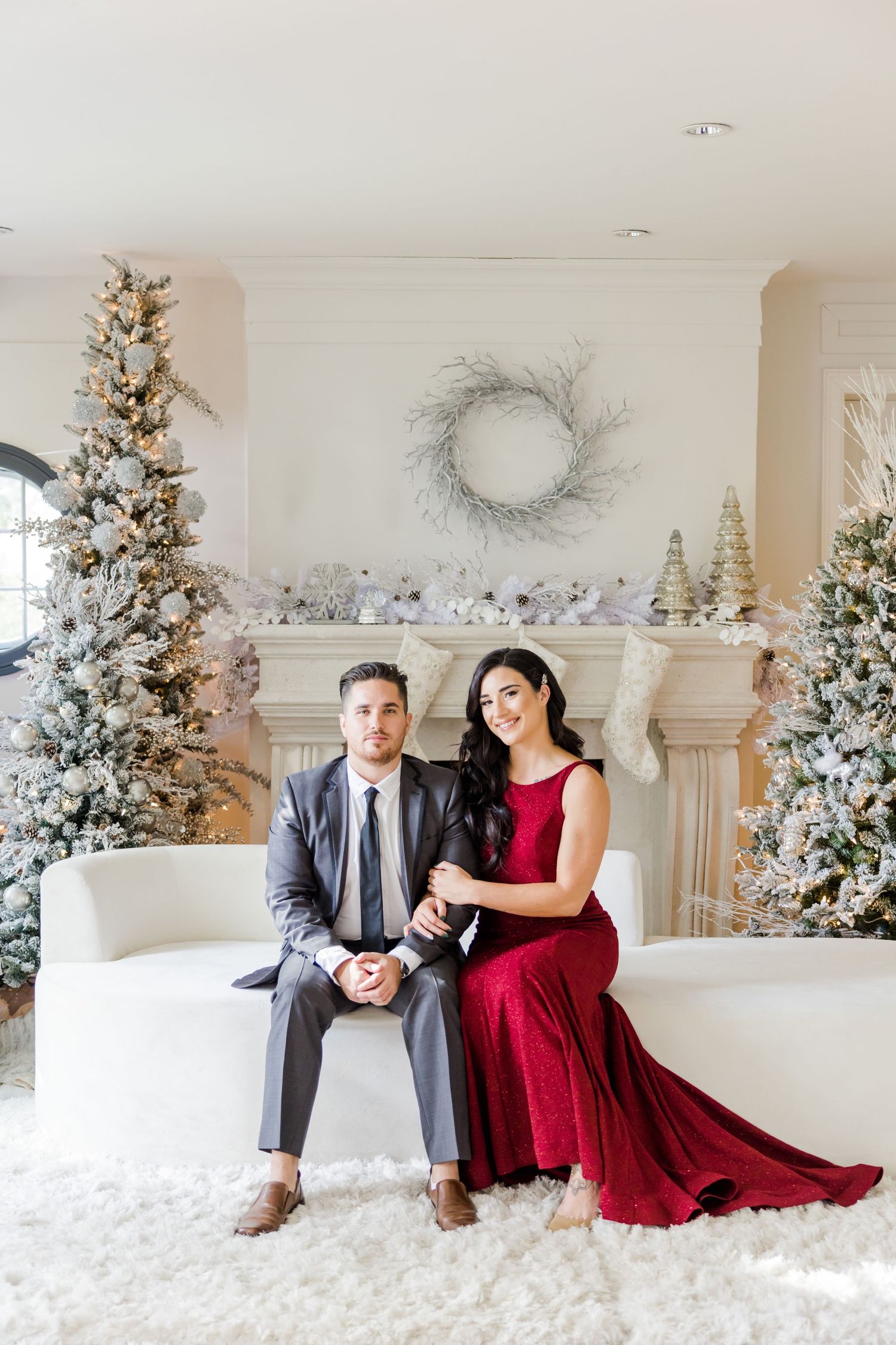 Holiday couple portrait with red gown and grey suit against festive Christmas backdrop.