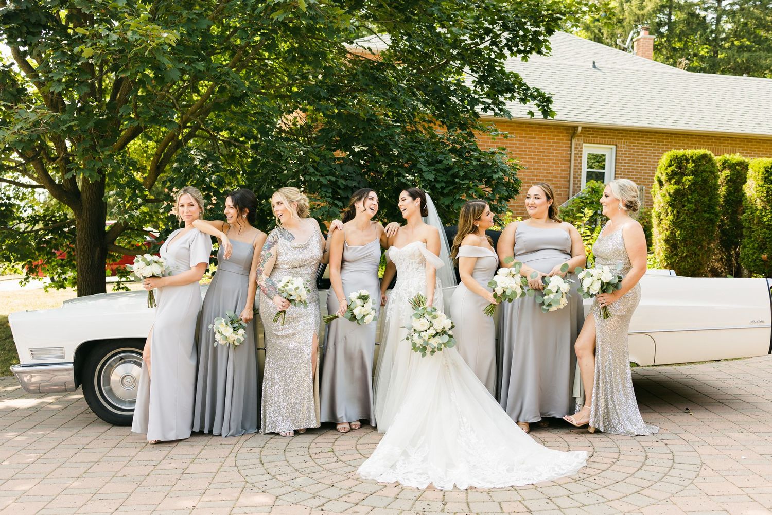 Bridesmaids in flowing grey dresses pose beside a classic white car at an outdoor wedding venue.