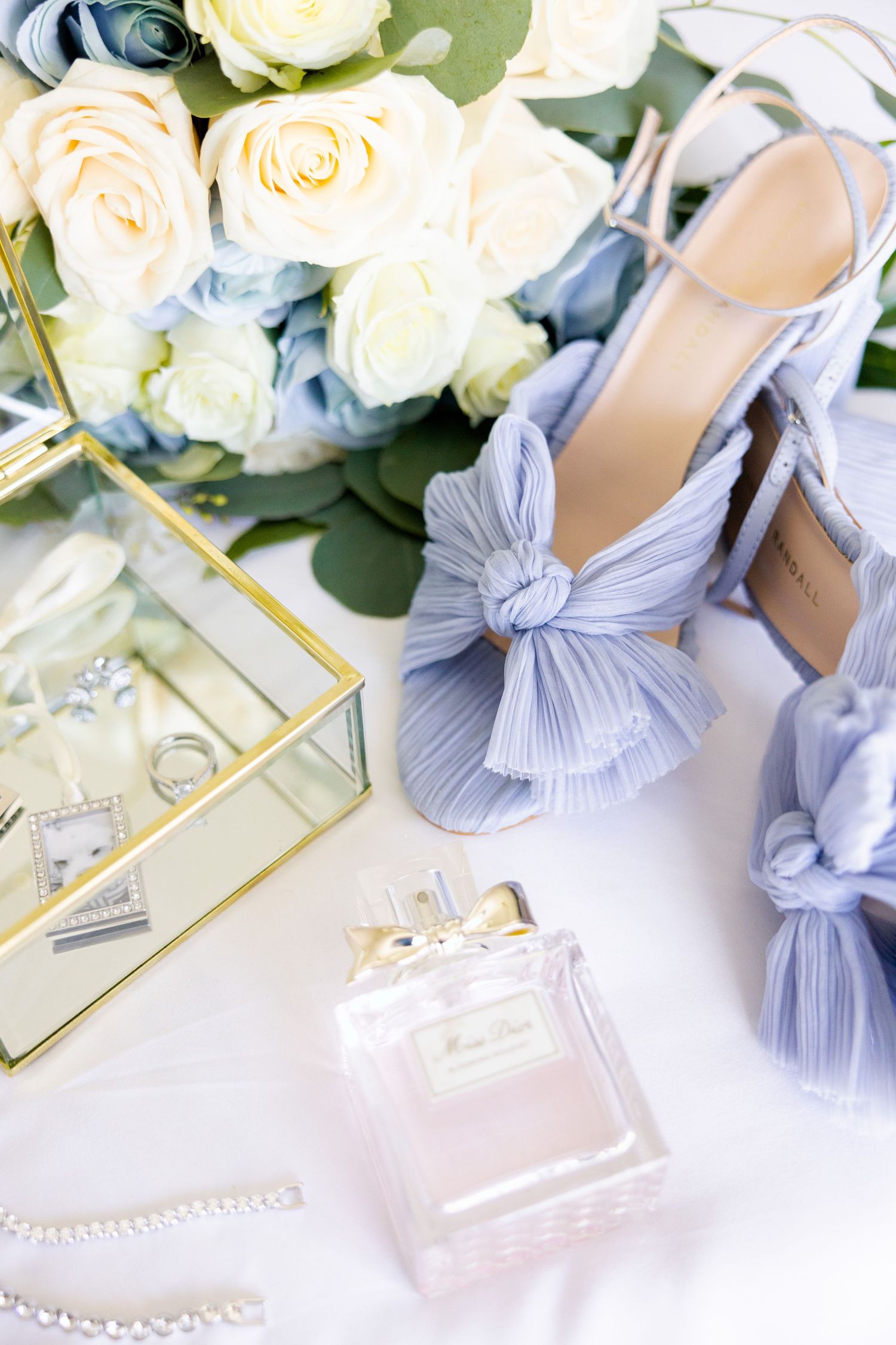 Elegant wedding accessories including cream roses, light blue bow heels, perfume bottle and glass jewelry box.