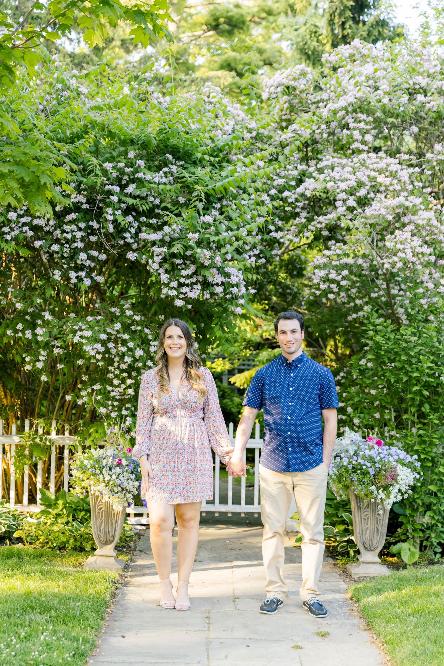 Couple stands on garden path with white flowering bushes and picket fence in background.