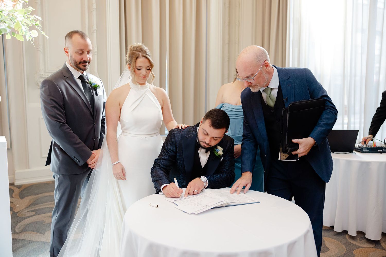 Wedding ceremony guests watching as a couple signs documents at an elegant white-decorated venue.