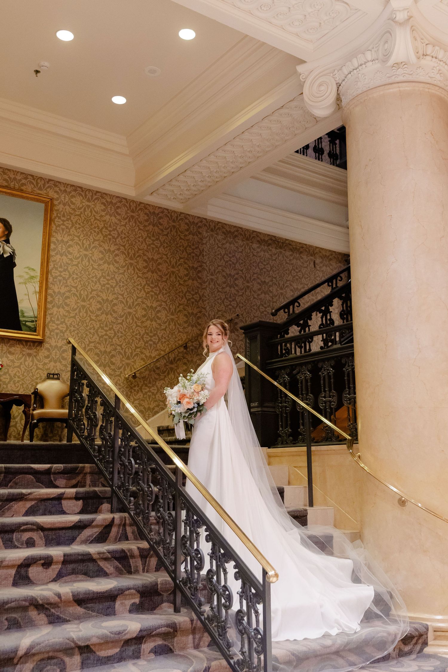 Elegant staircase with ornate iron railings and columns in a grand venue features flowing white wedding dress train.