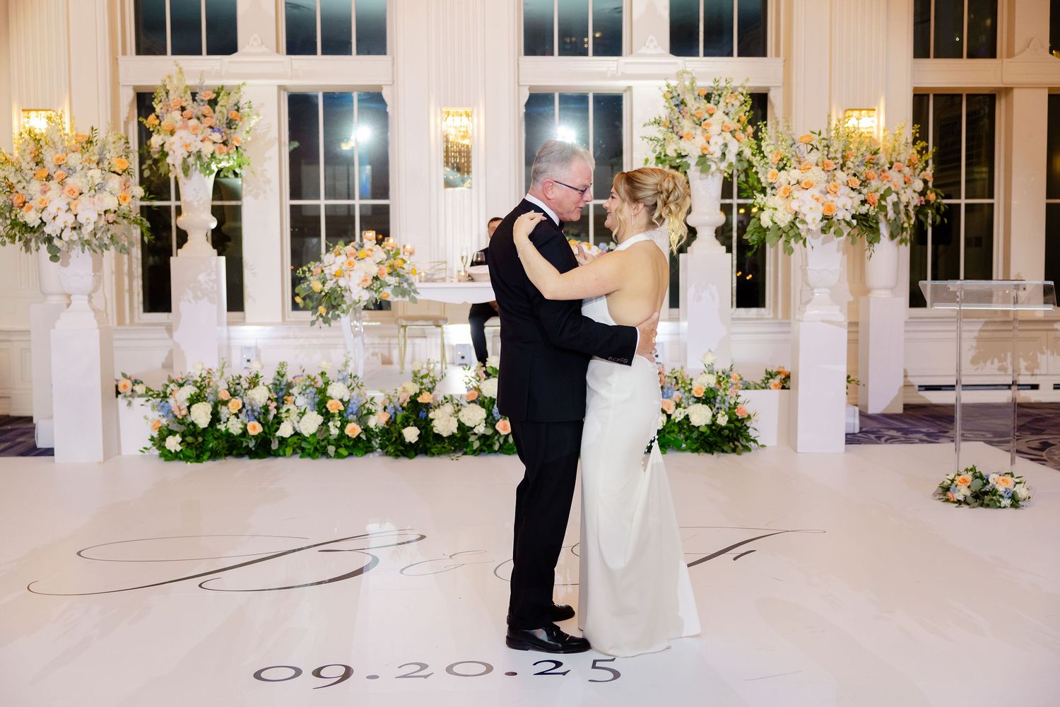 Romantic first dance in an elegant white room decorated with floral arrangements and tall windows.