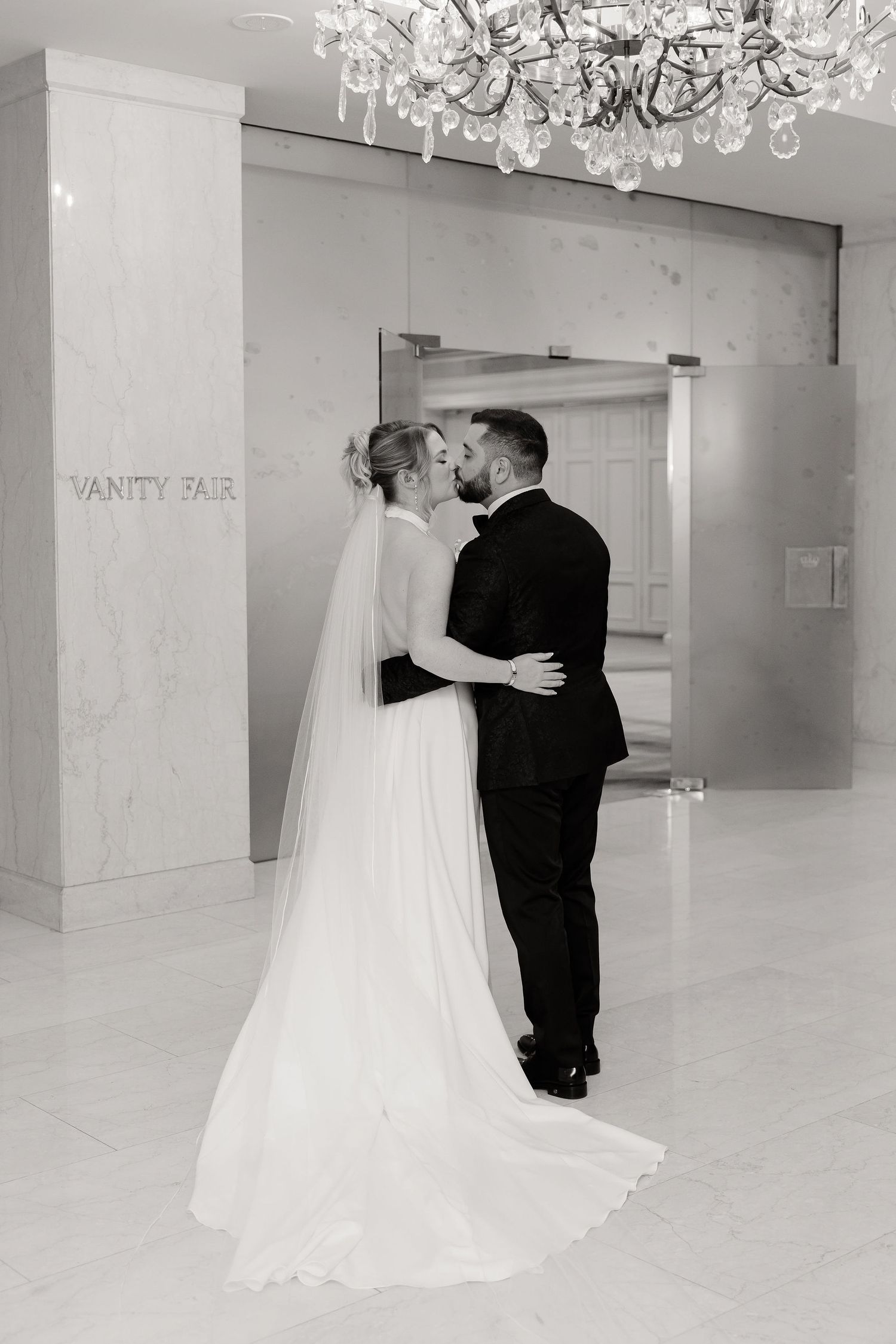 Black and white wedding photos captured in a modern white ballroom with crystal chandeliers.