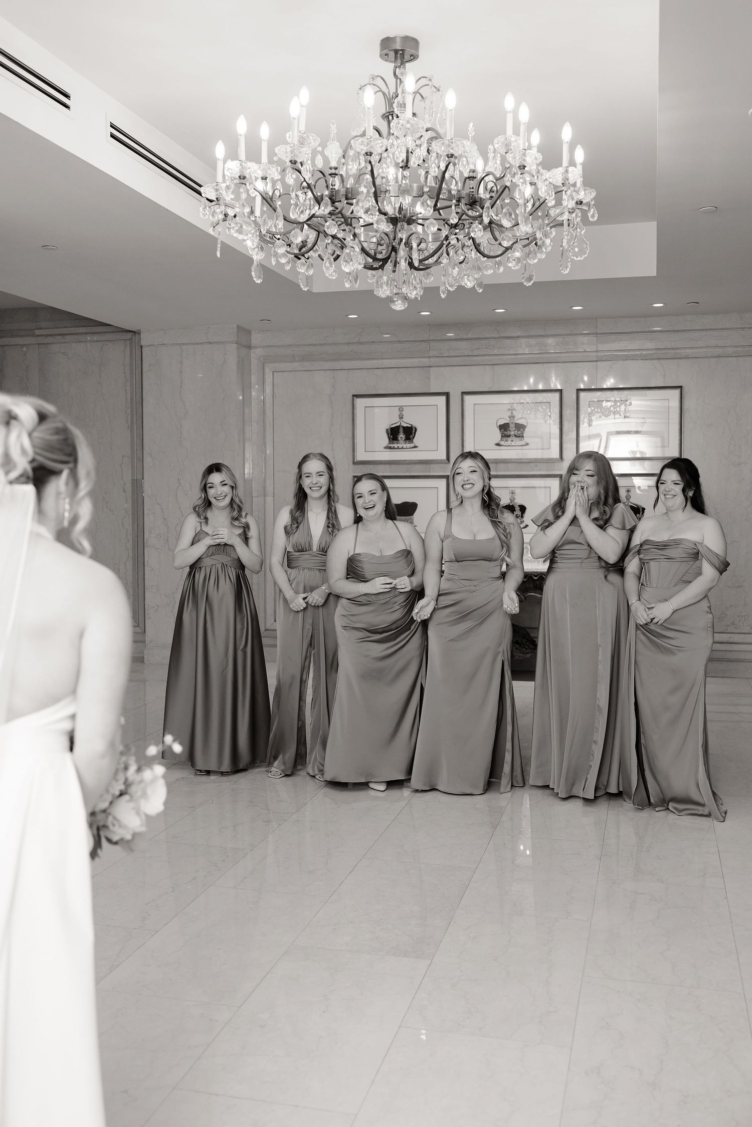 Bridesmaids in elegant gray dresses pose in a luxurious ballroom with crystal chandeliers.