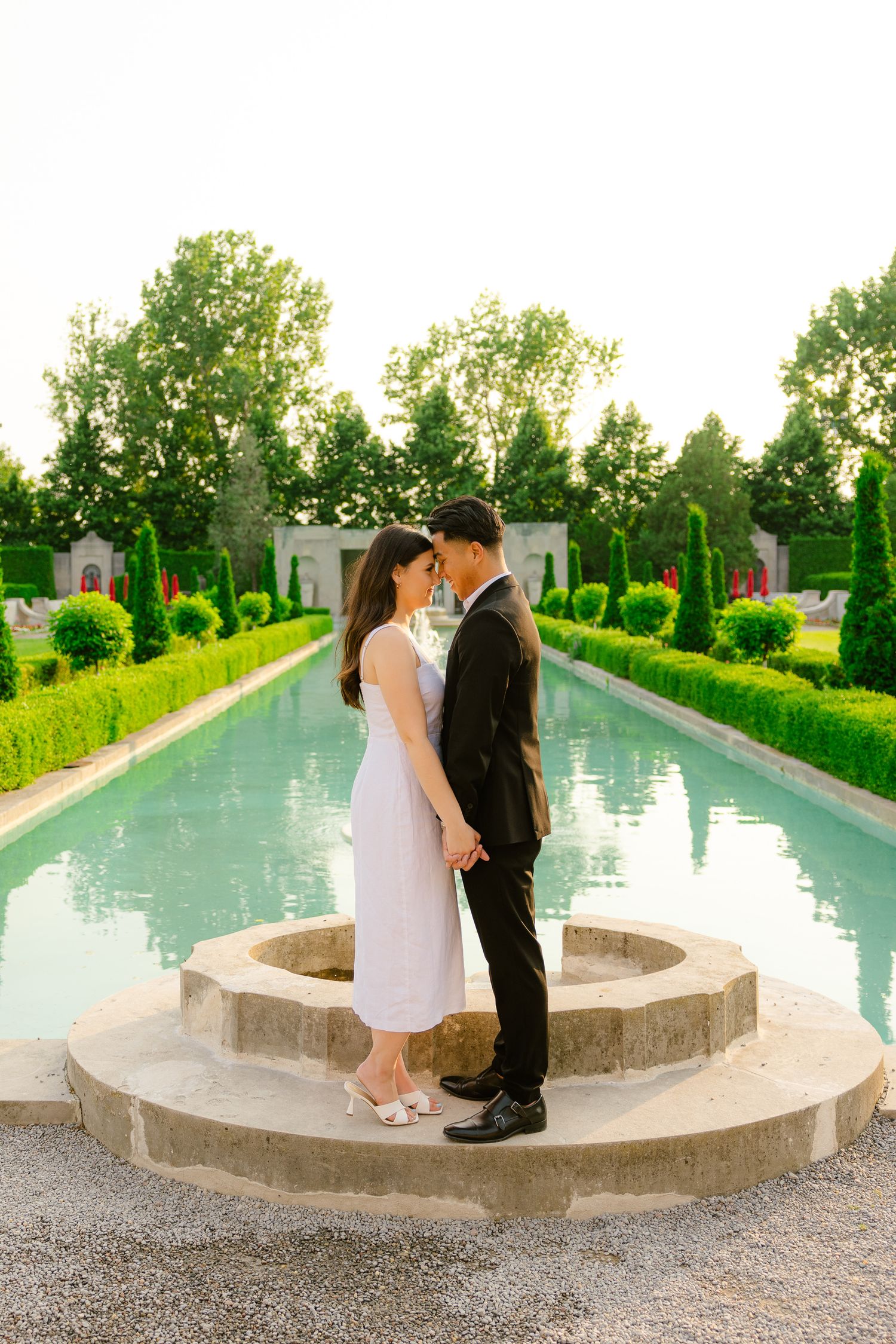 A couple embracing on circular stone steps with a reflecting pool and manicured garden in the background.