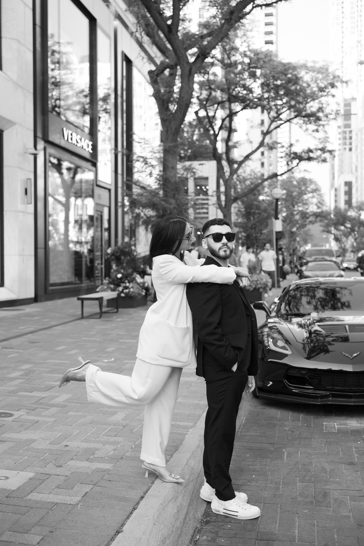 Black and white photo of couple in contrasting suits embracing playfully on urban sidewalk.