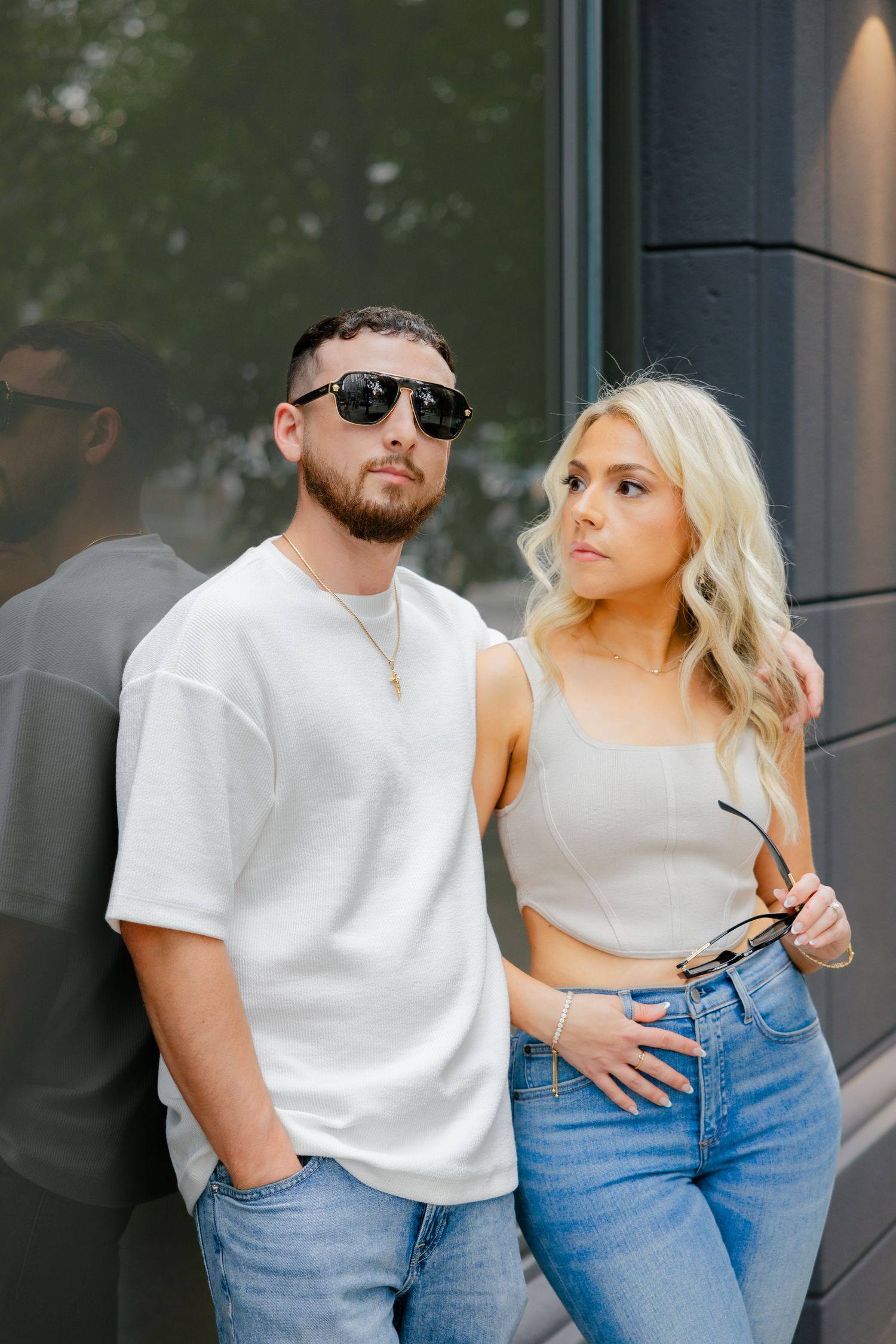 A couple in casual attire posing against a modern building exterior.