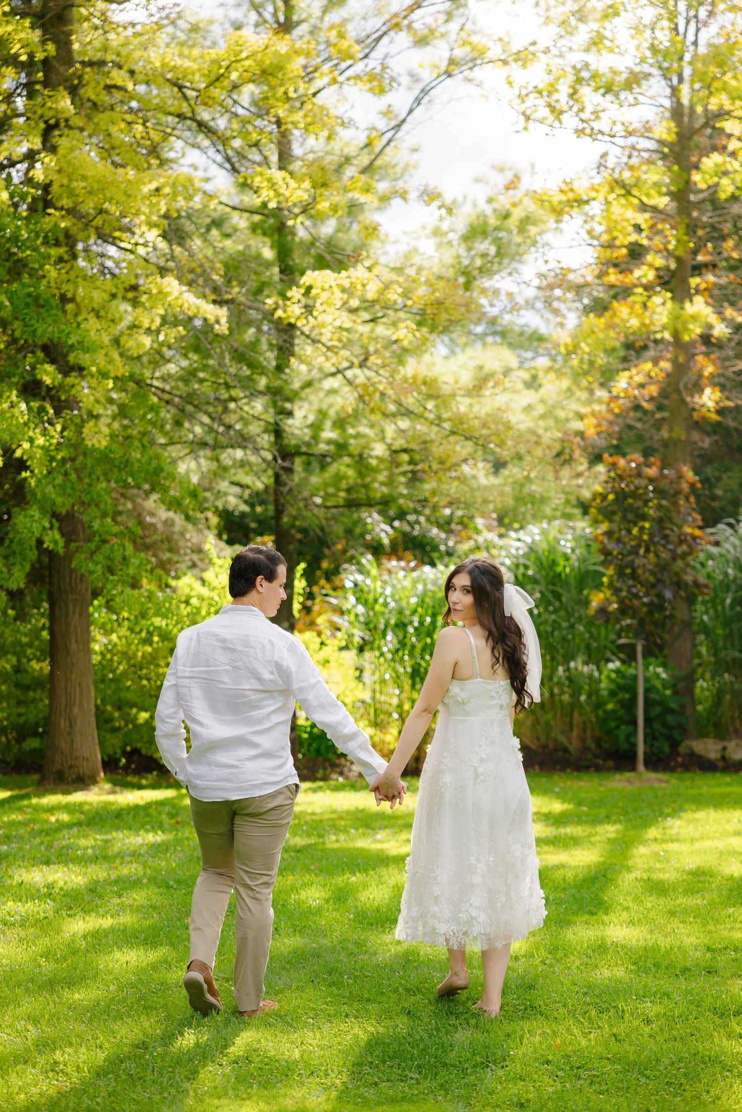 Couple in wedding attire walking hand in hand through a sunlit garden with trees.