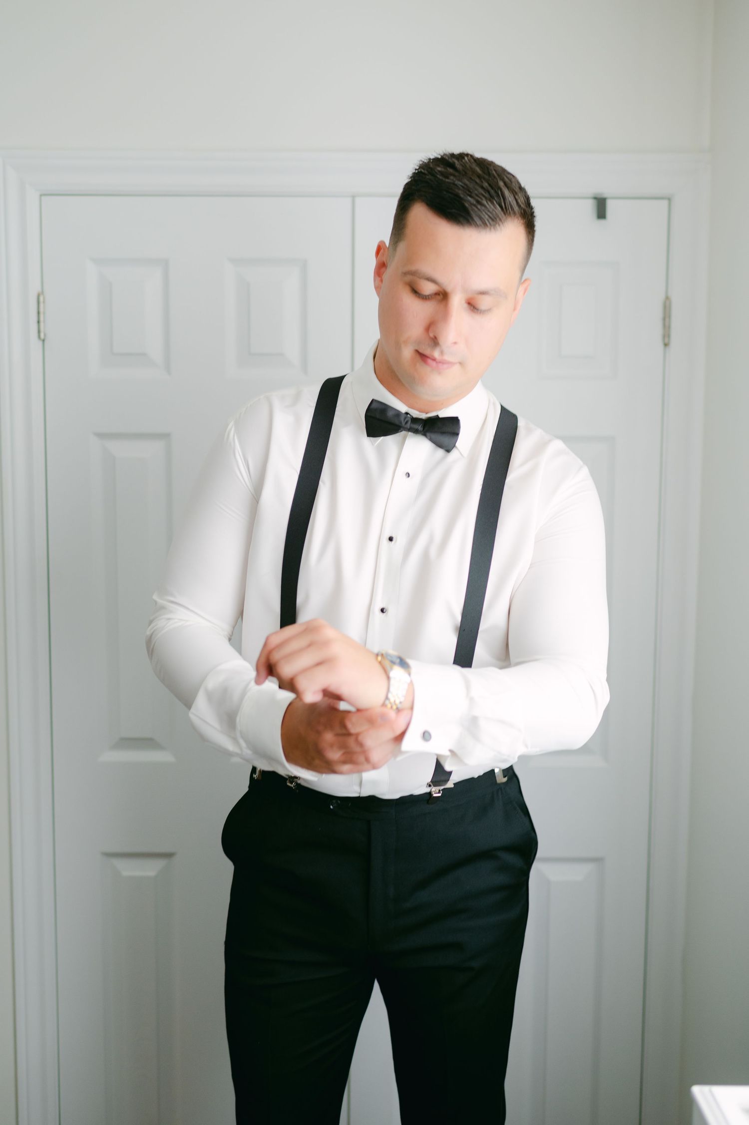 A formal portrait of someone adjusting their black bow tie while wearing suspenders and a white dress shirt.