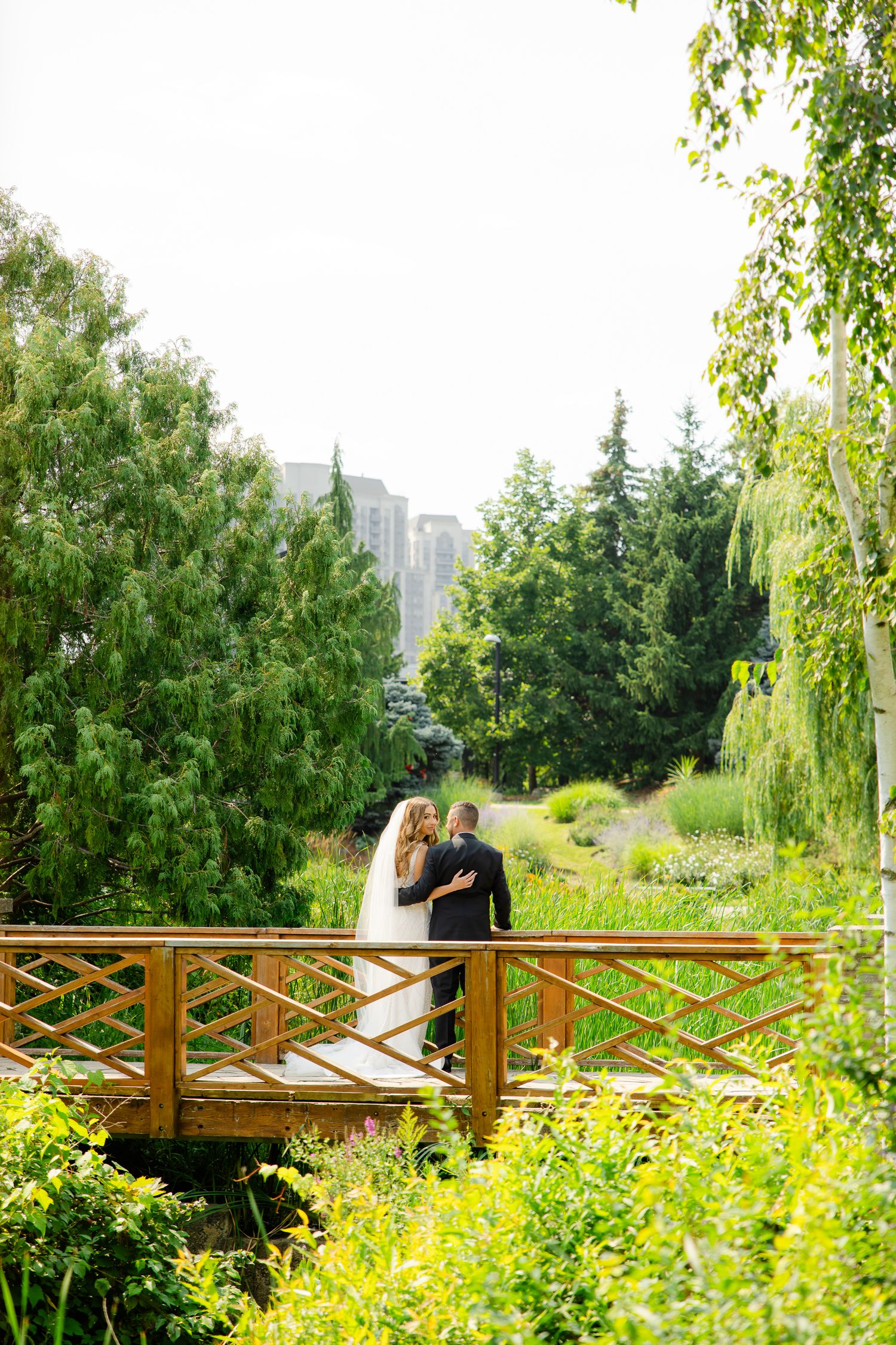 Romantic wedding couple standing on a wooden bridge surrounded by lush garden scenery and tall trees.
