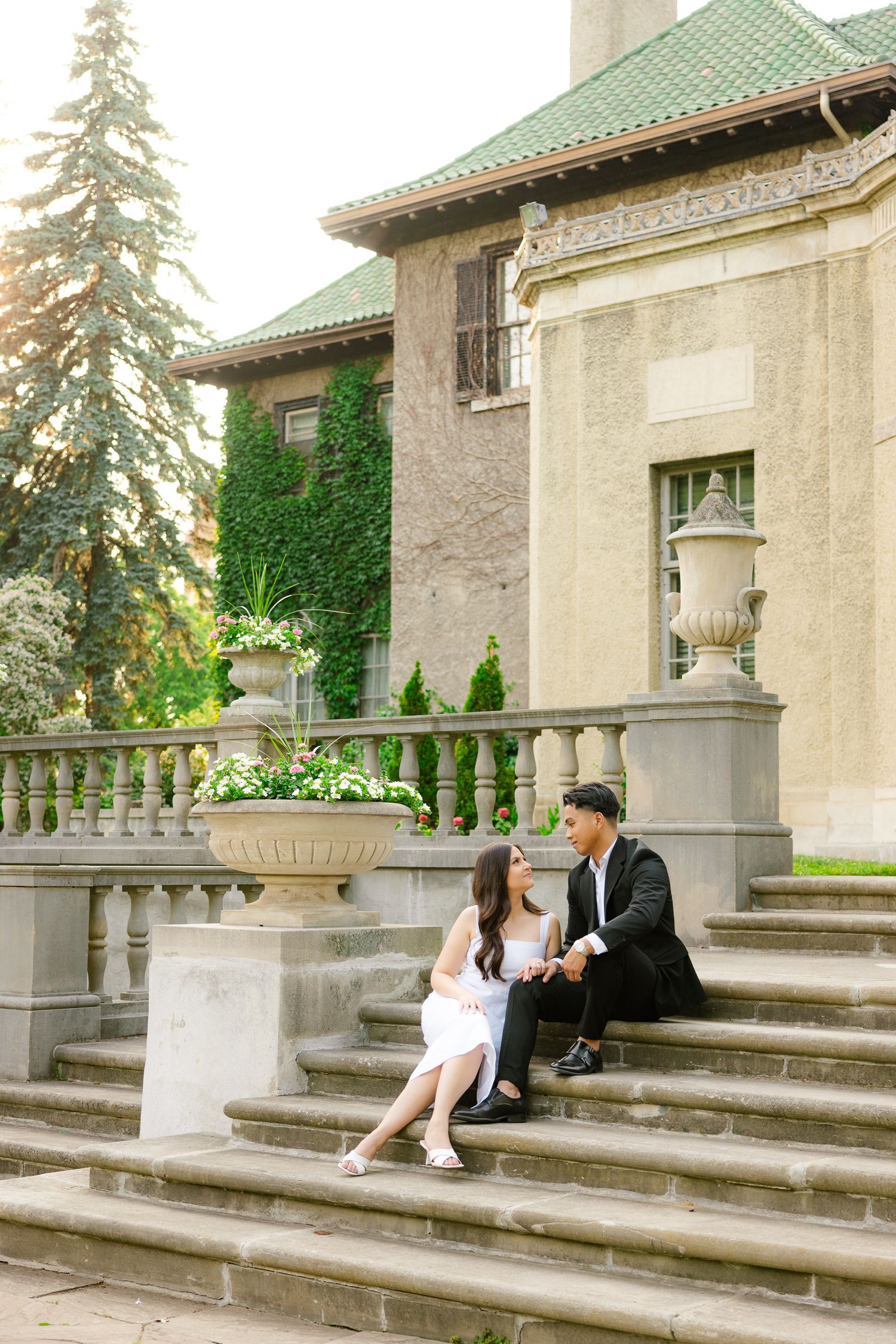 Couple sits on steps of an elegant stone building with ivy-covered walls and green roof.