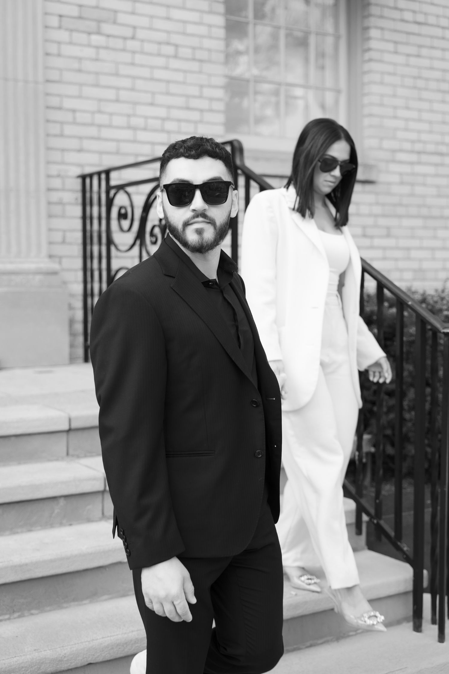 Elegant couple in sunglasses and contrasting suits stand on steps of brick building.