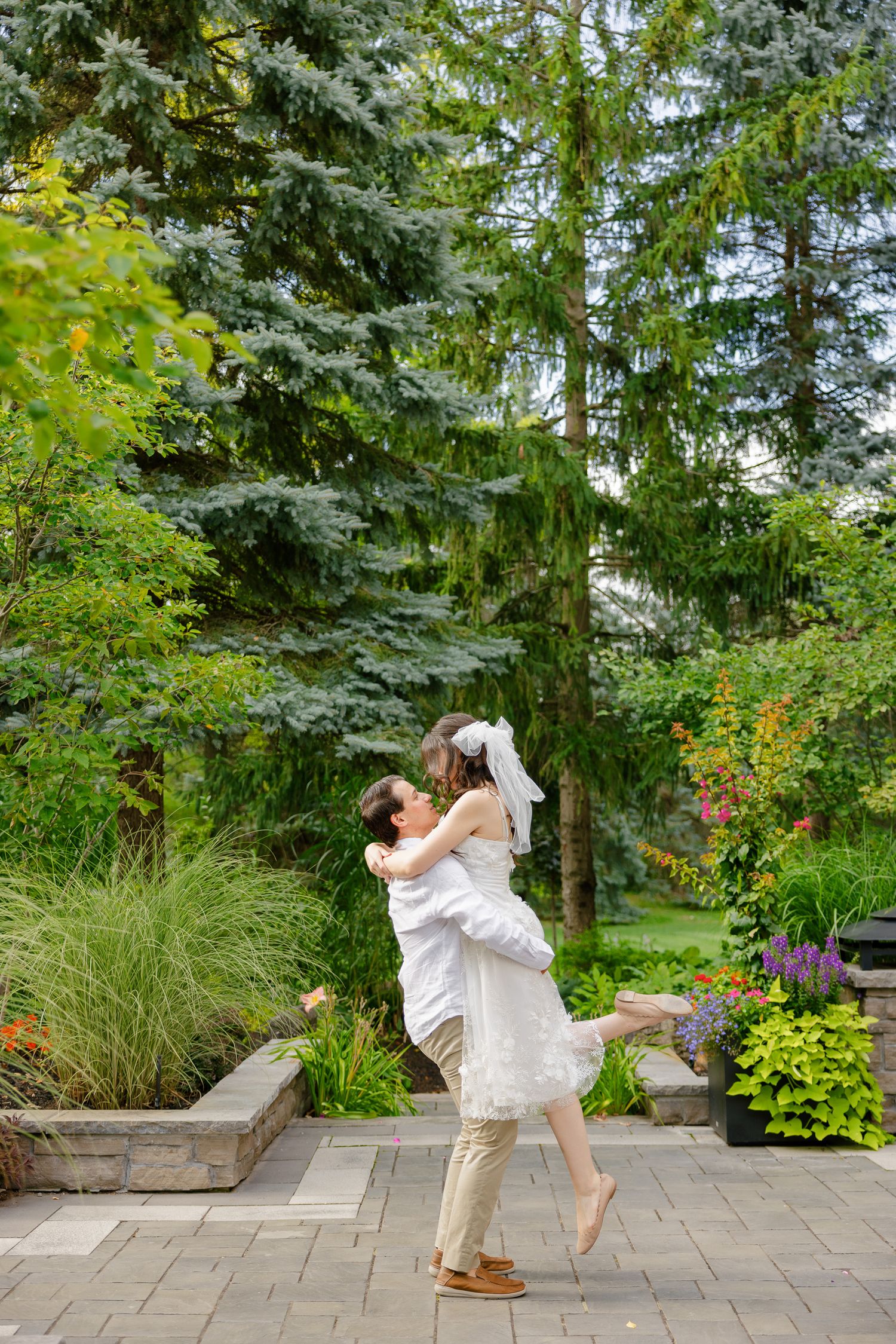 A romantic moment captured in a garden as a couple embraces and spins on a stone patio surrounded by evergreen trees.