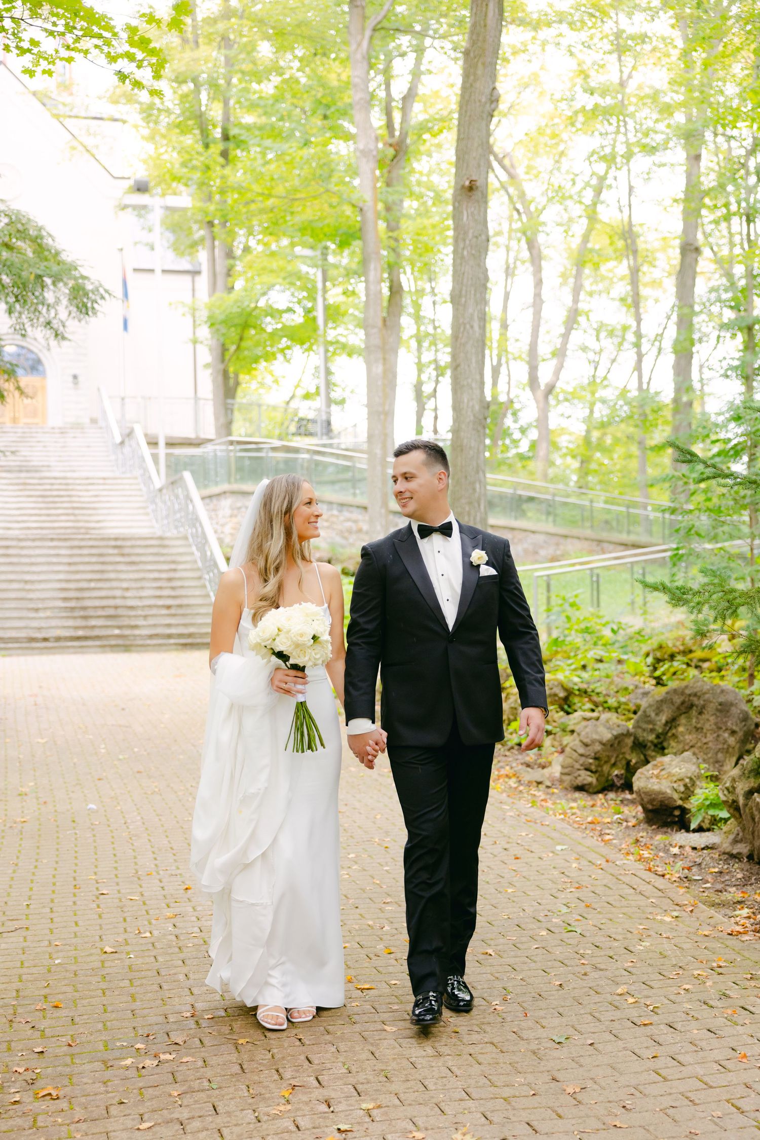 Bride and groom walking hand in hand along brick pathway surrounded by trees.