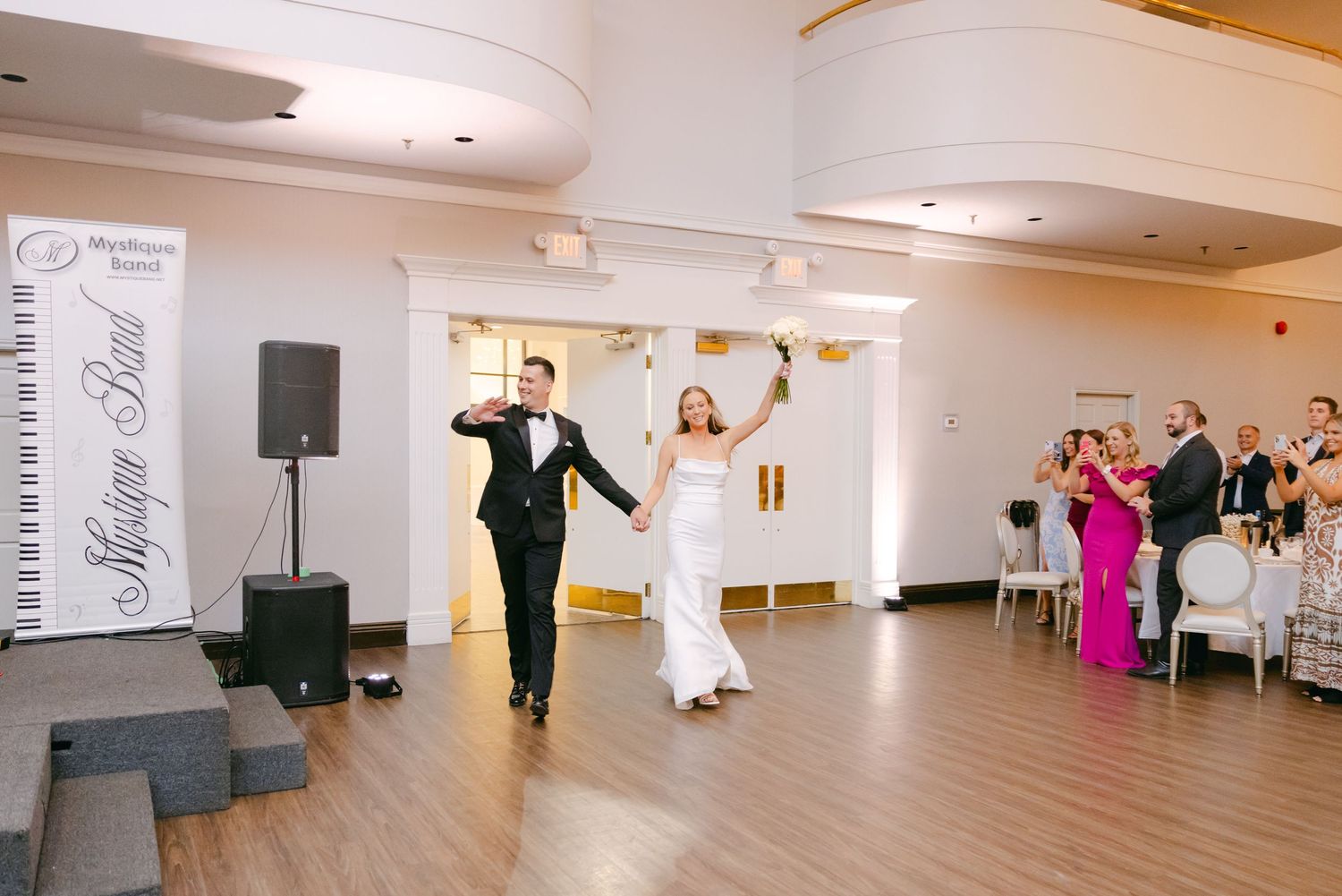 Wedding couple makes grand entrance into reception hall decorated with white draping and wooden floors.