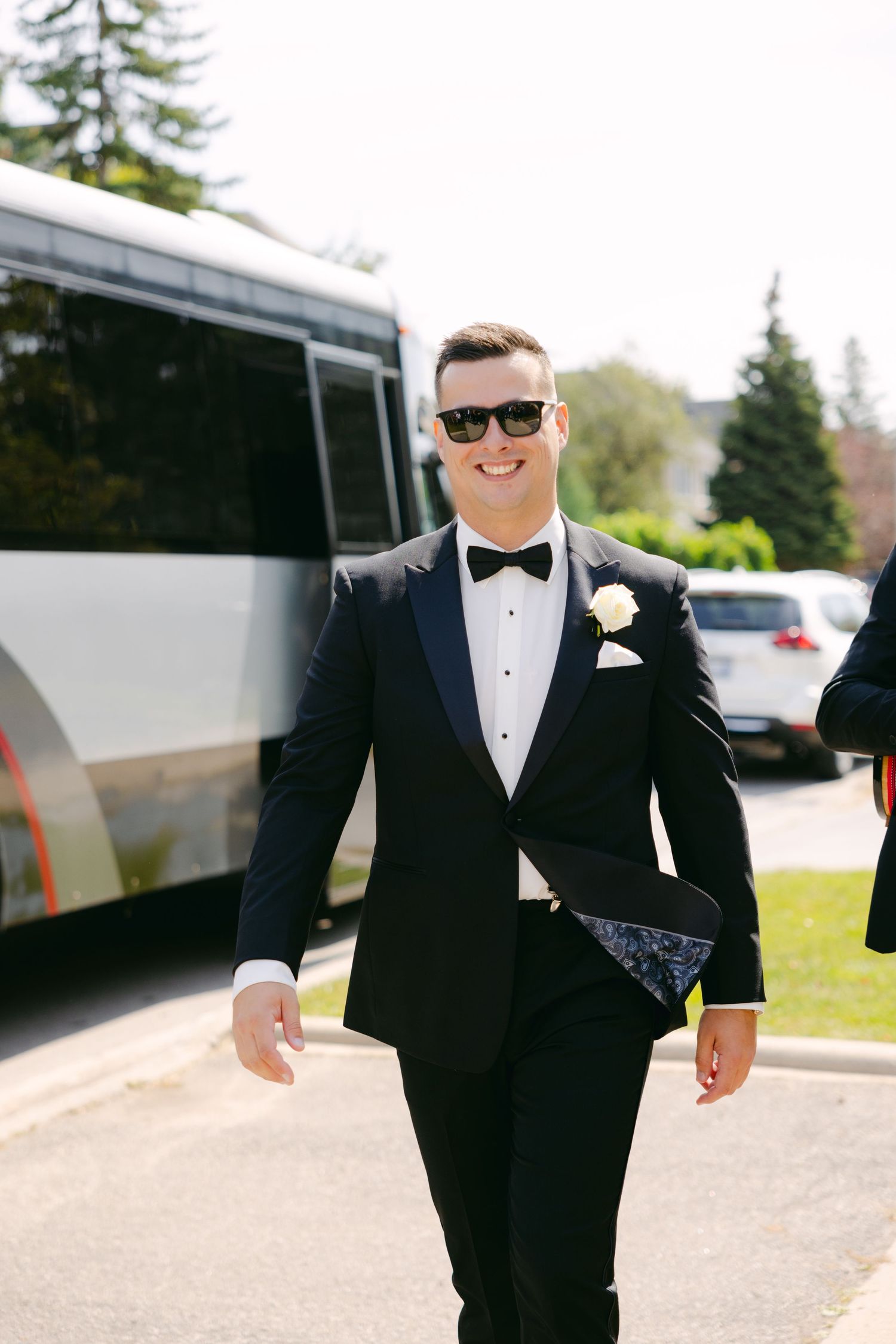 Groom in classic tuxedo and sunglasses smiles while walking outside.