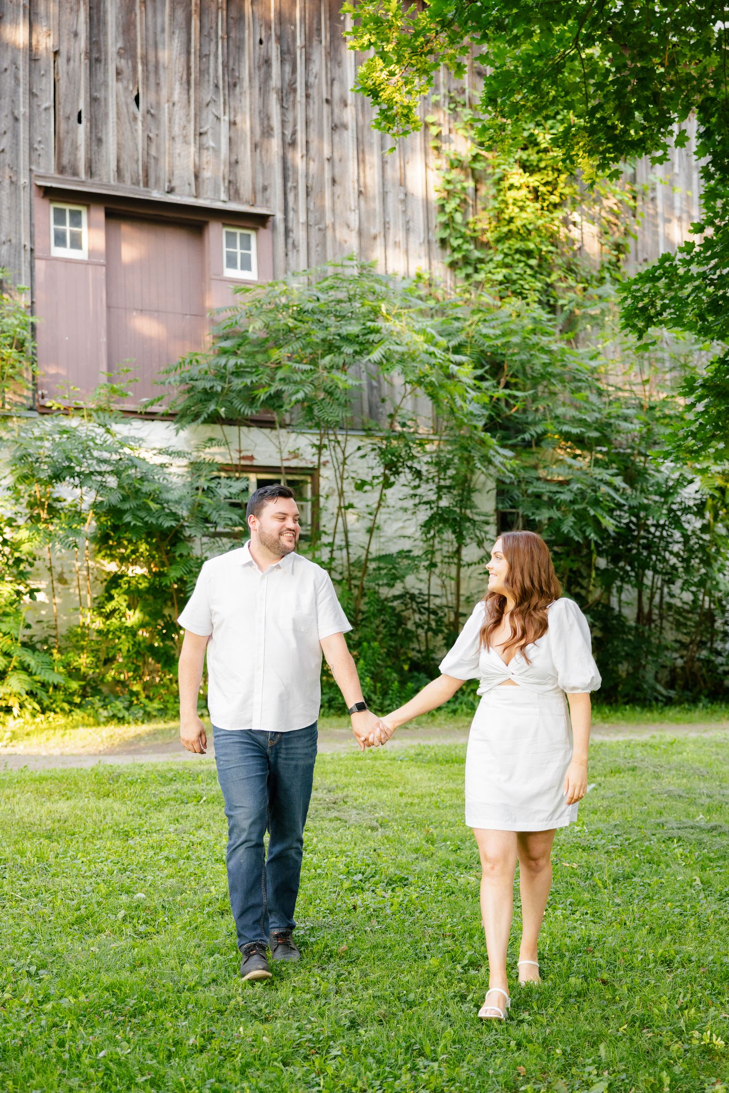 Couple in white holding hands while walking in front of a rustic barn on a sunny day.
