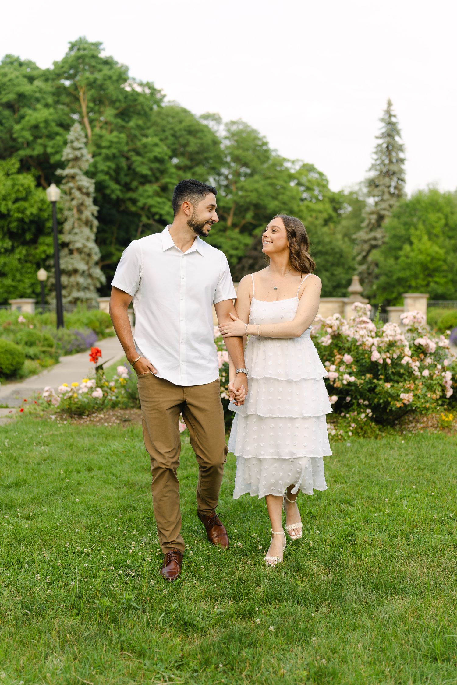 A couple strolls together through a lush garden setting with blooming roses and evergreen trees.