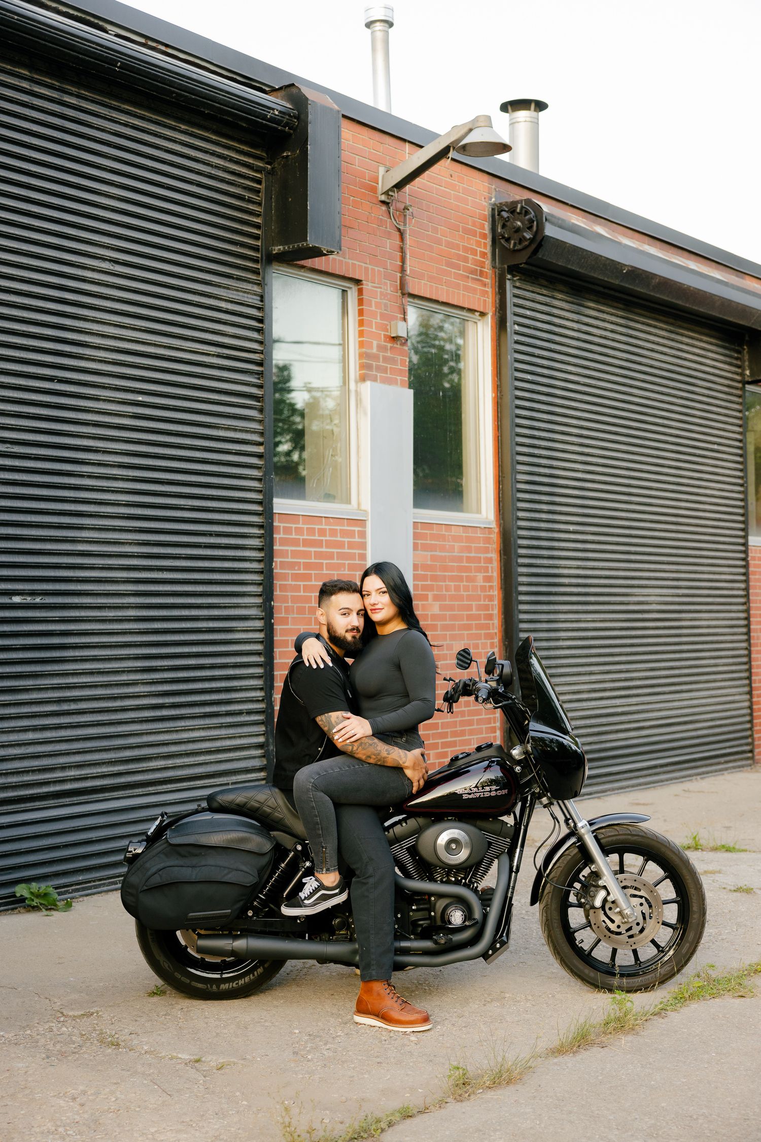 A couple cuddles on a Harley Davidson motorcycle against an industrial backdrop.