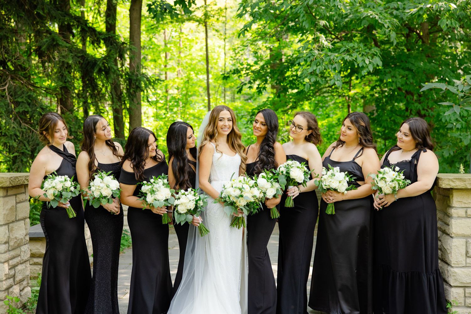 Wedding party with bridesmaids in black dresses holding white bouquets pose against lush green garden backdrop.
