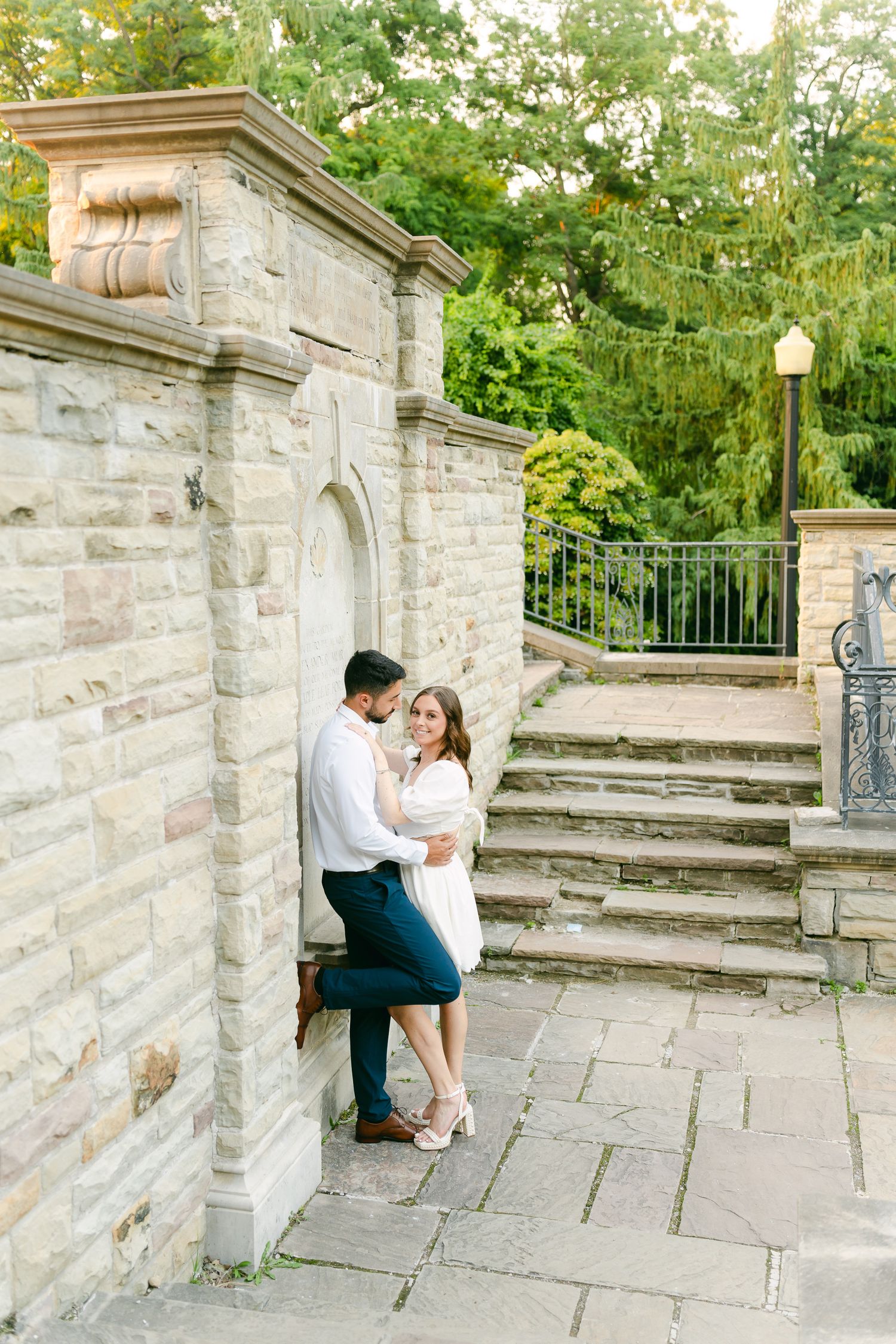 A couple embraces near stone steps with lush greenery and a lamp post in the background.
