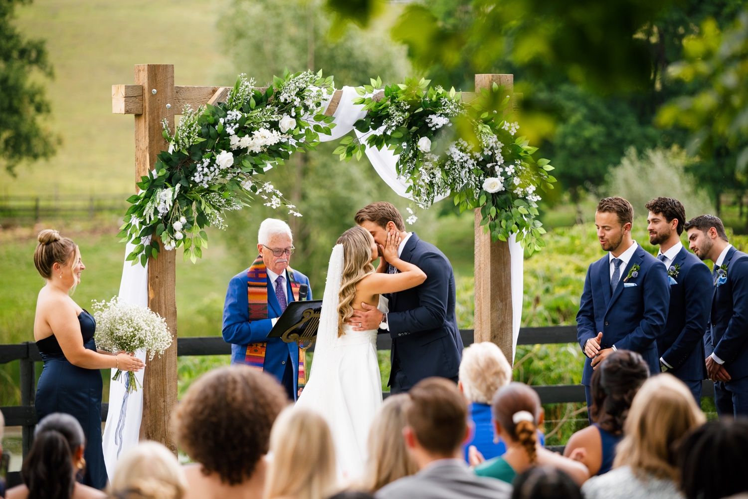 Wedding guests observe a ceremony under a floral-decorated wooden cross arch in an outdoor setting.