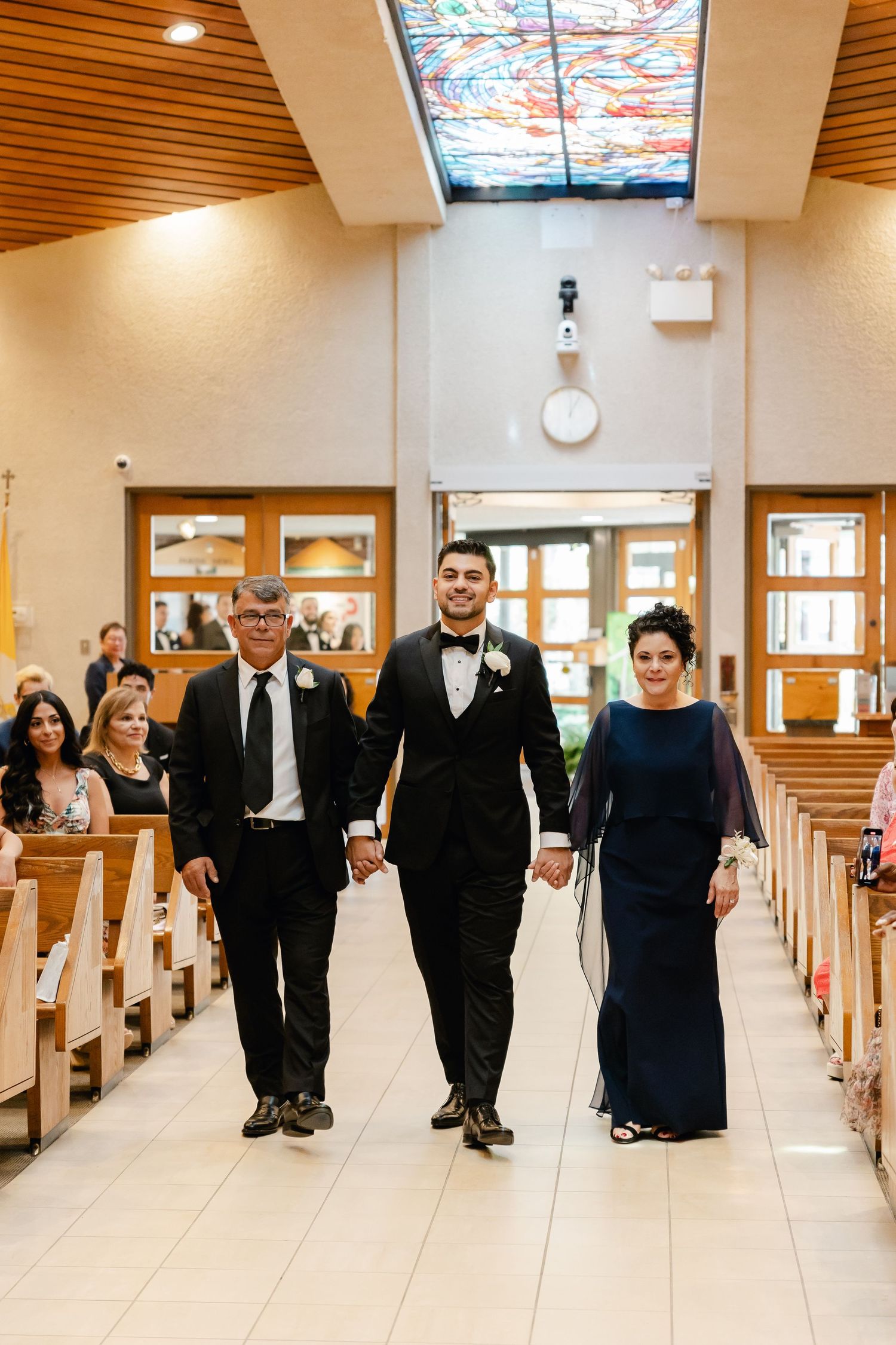 Wedding processional with family members walking down church aisle against ornate stained glass skylight ceiling.