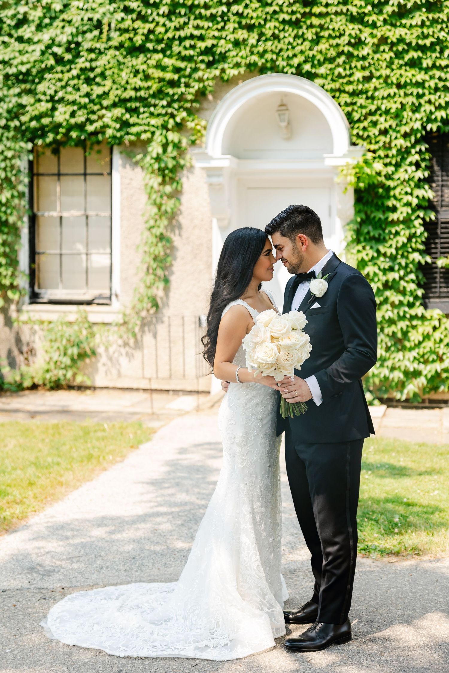 Newlyweds standing on a garden path with white flowers and ivy-covered walls behind them.
