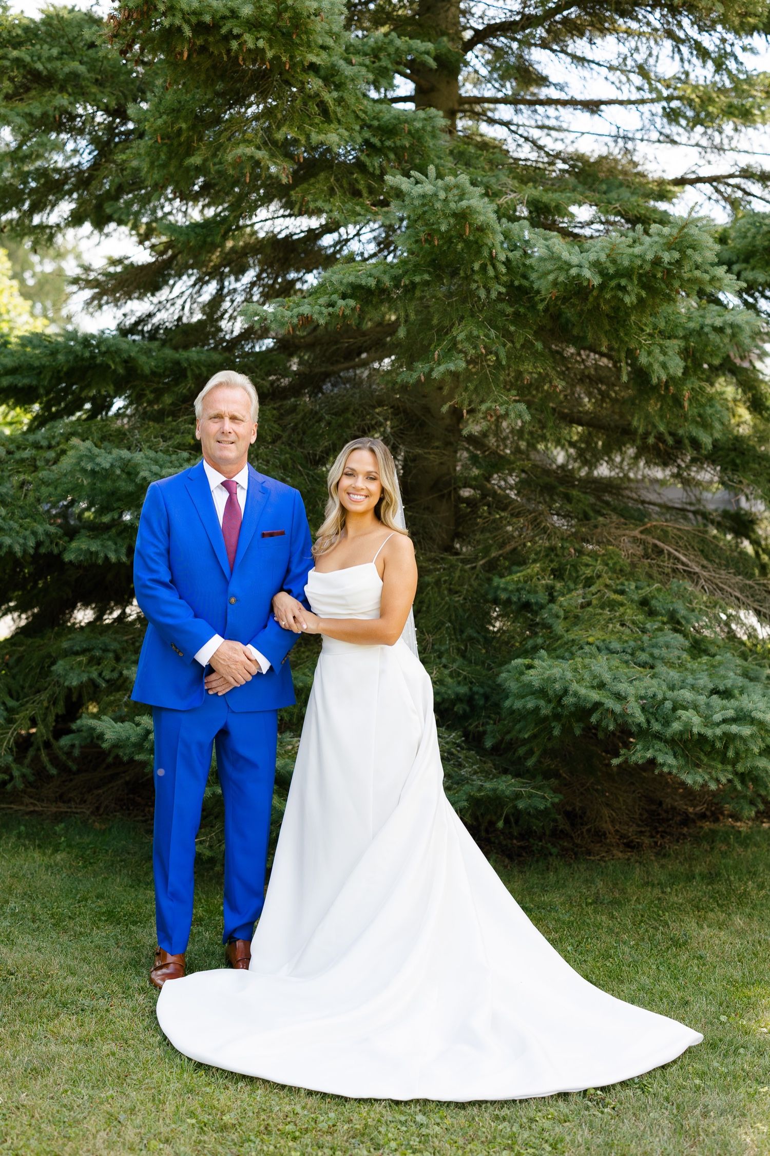 Outdoor wedding portrait against evergreen trees featuring bright blue and white attire.
