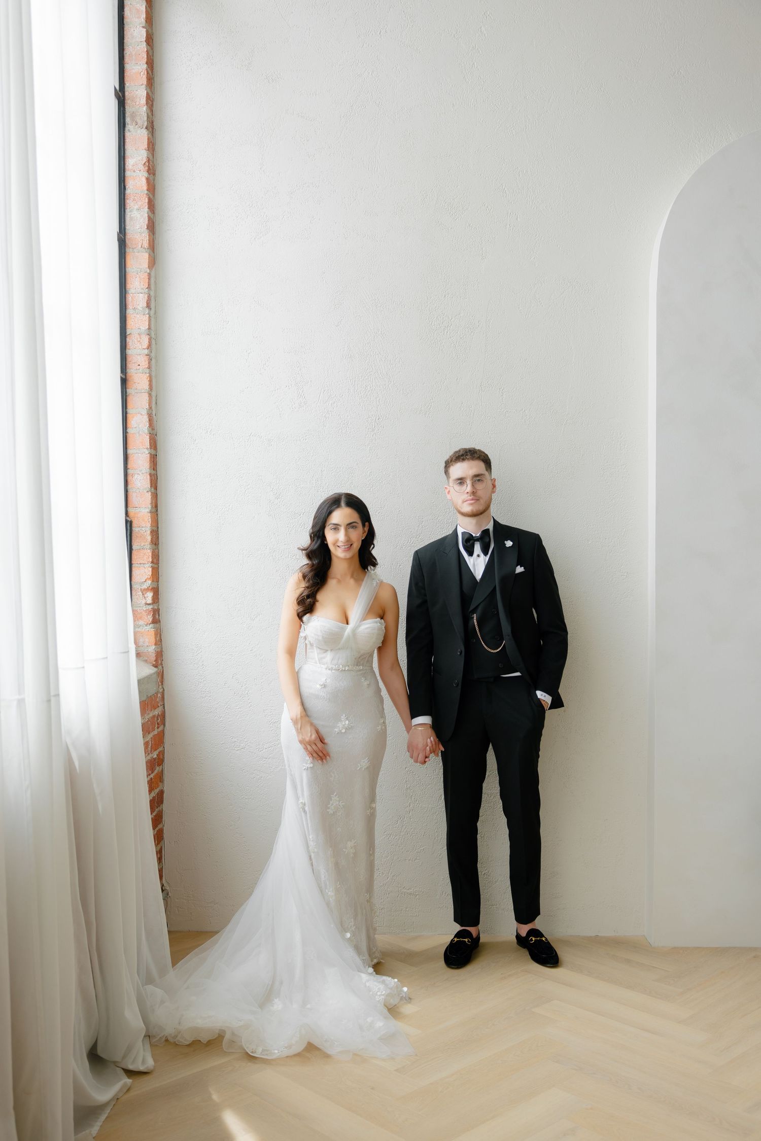 Elegant wedding portrait featuring a strapless lace gown and formal black tuxedo against white walls.