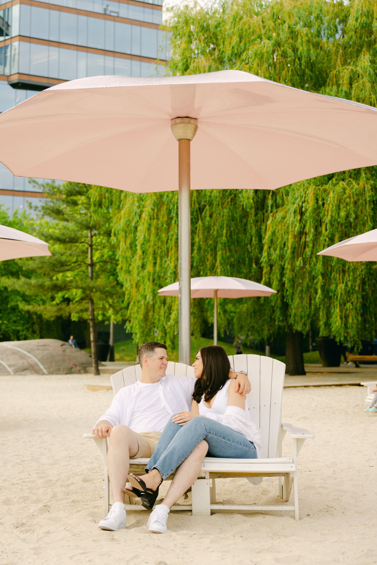 A couple sits together on a white beach chair under pink umbrellas with willow trees in background.